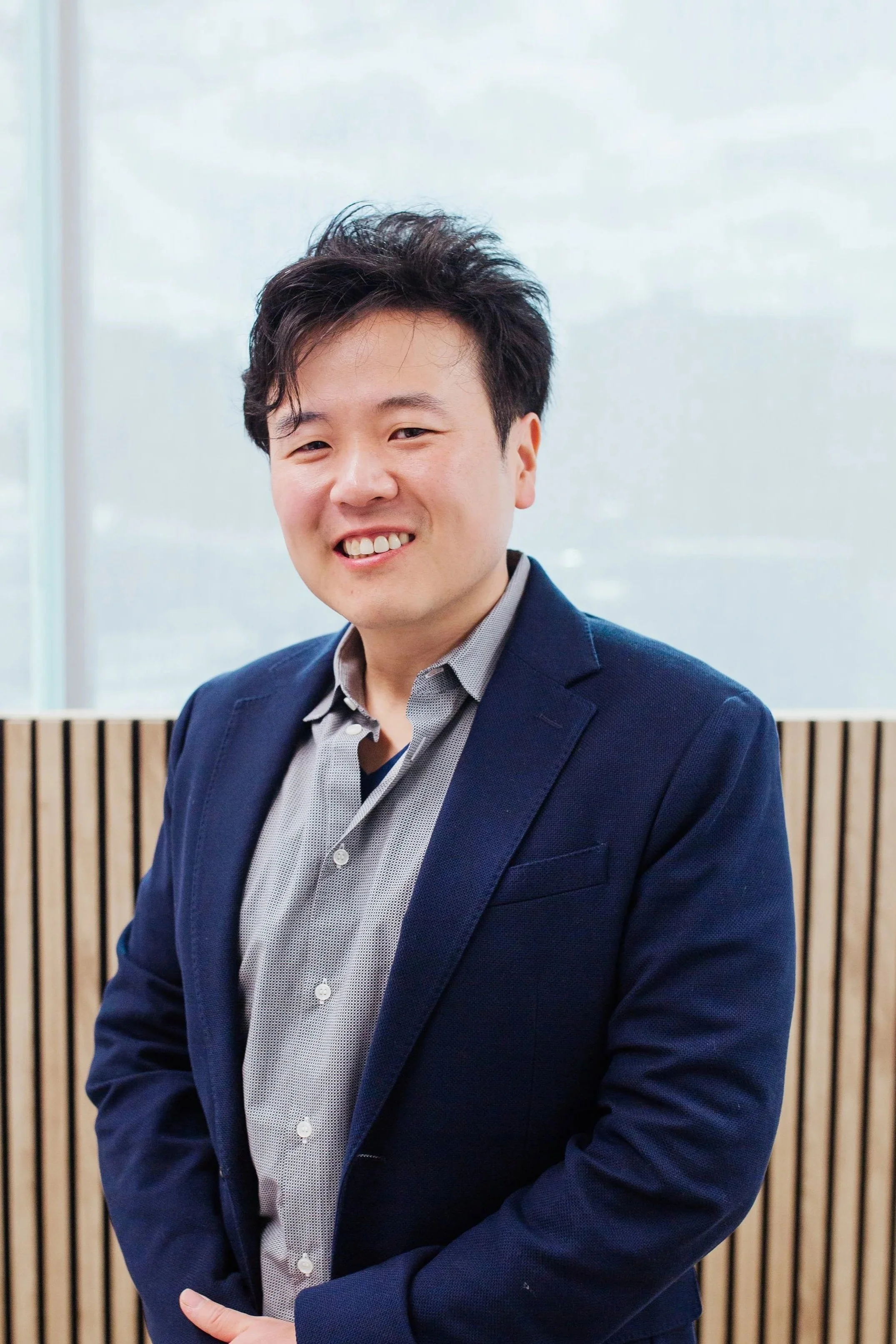 A young man with short, dark, textured hair smiling and wearing a dark blue blazer over a light gray patterned shirt, standing in front of a background with vertical wooden slats and a frosted glass window.