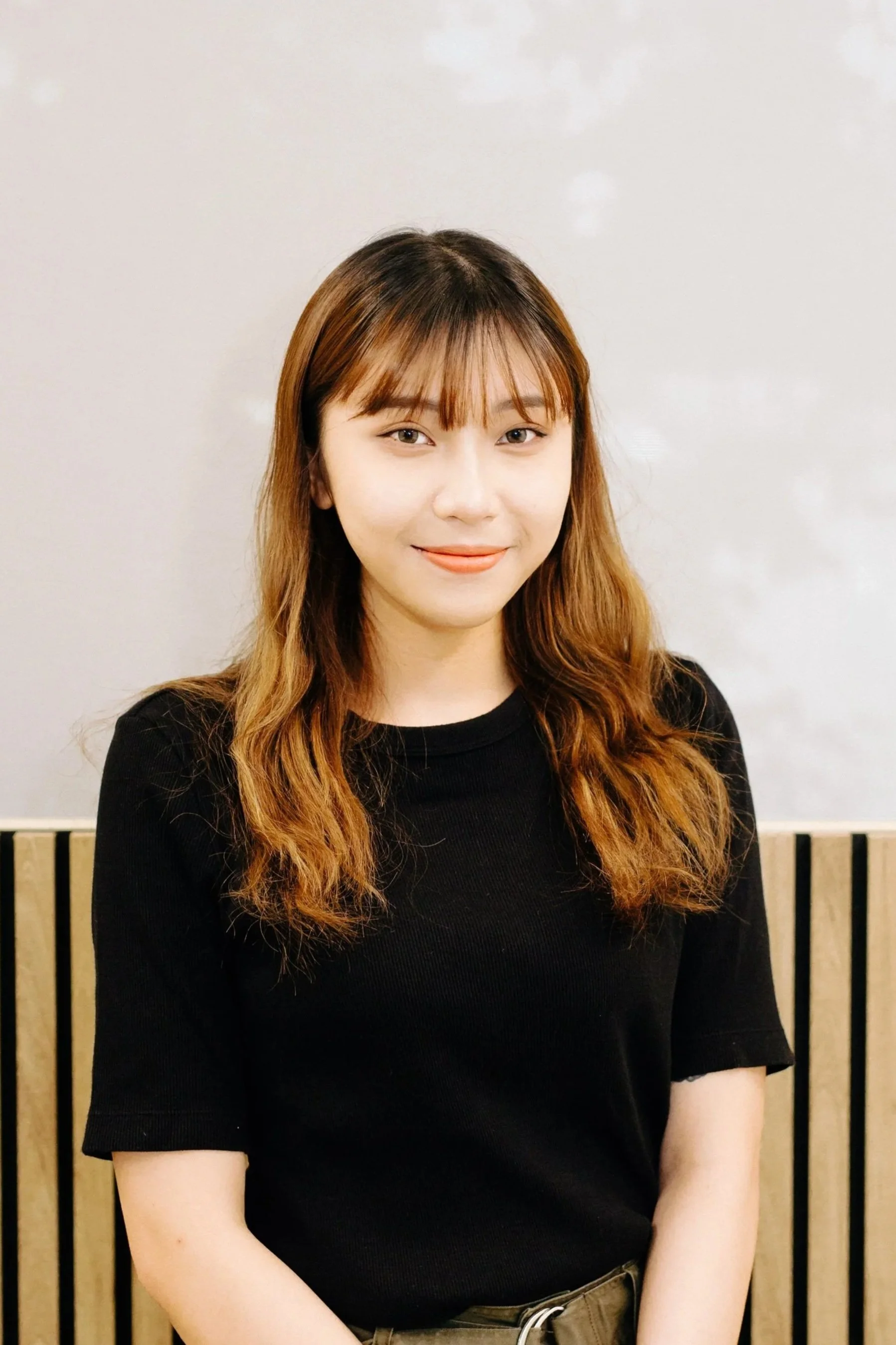 A young woman with brown hair and bangs, wearing a black shirt, standing in front of a wooden slatted wall.