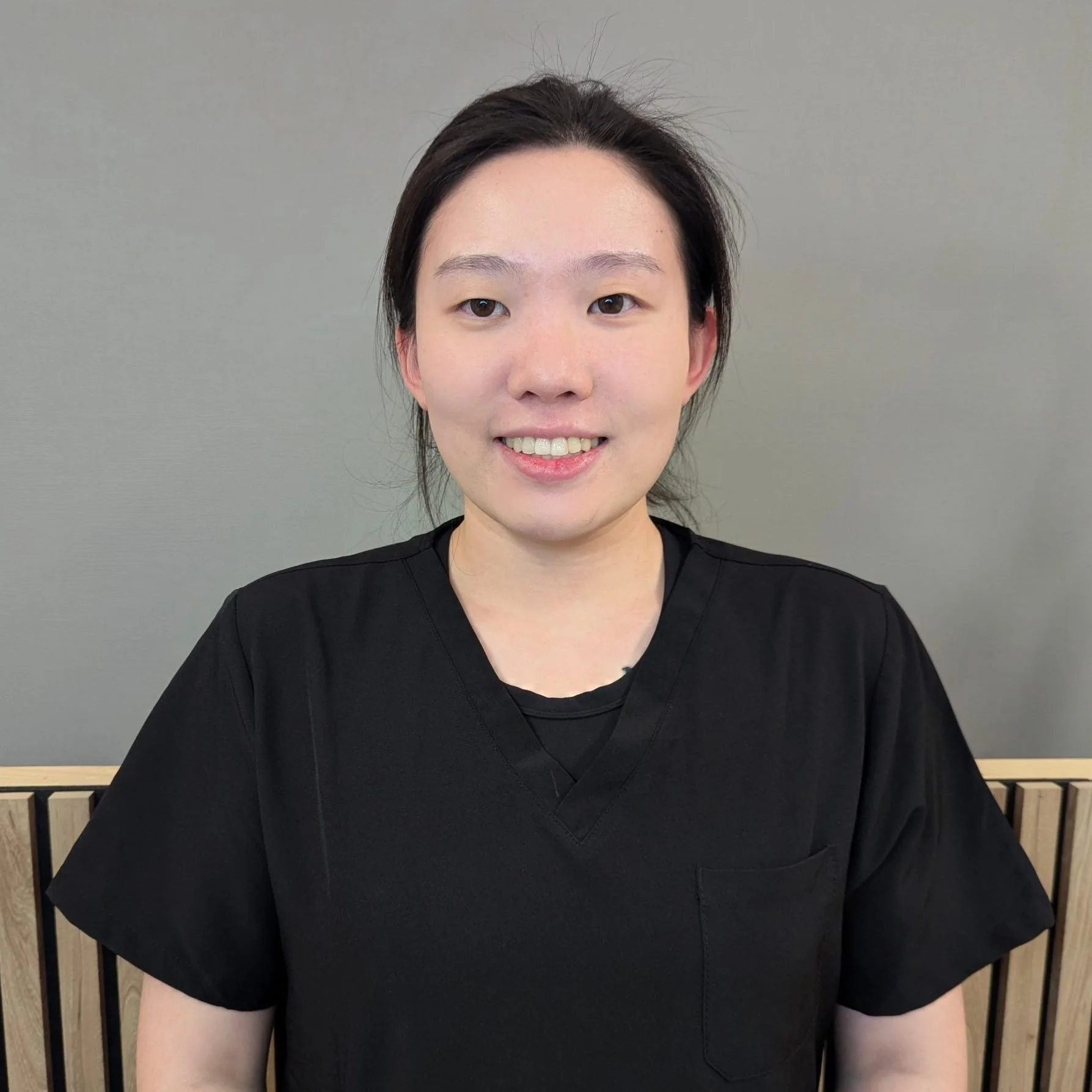 A smiling woman wearing black scrubs standing against a plain gray wall.