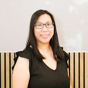 A woman with glasses and long hair smiling while standing indoors against a wall with round decorative wall hangings, wearing a white blouse.