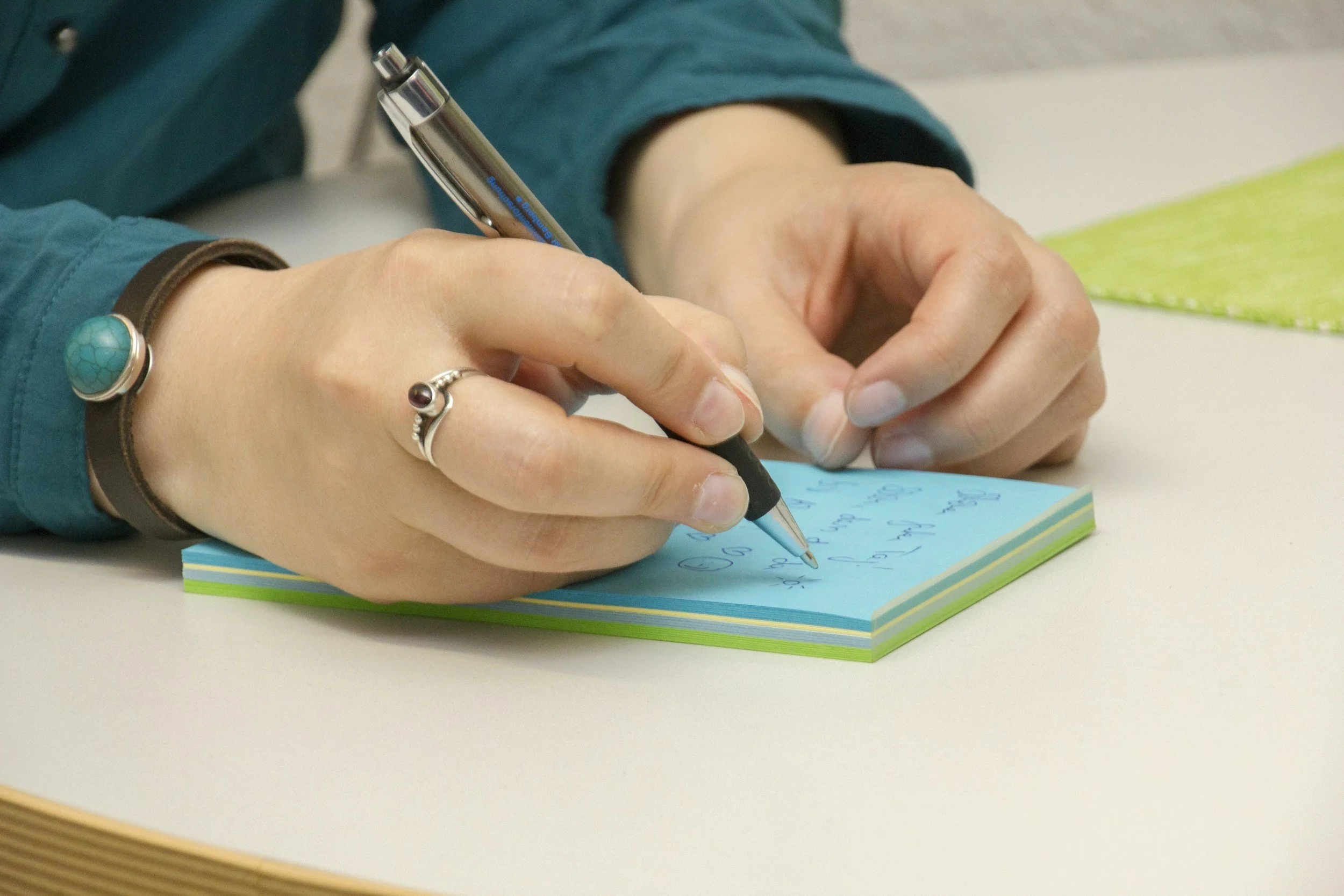 Person writing on a small blue notepad with a black pen, wearing a turquoise bracelet with a large turquoise ring, sitting at a white table.