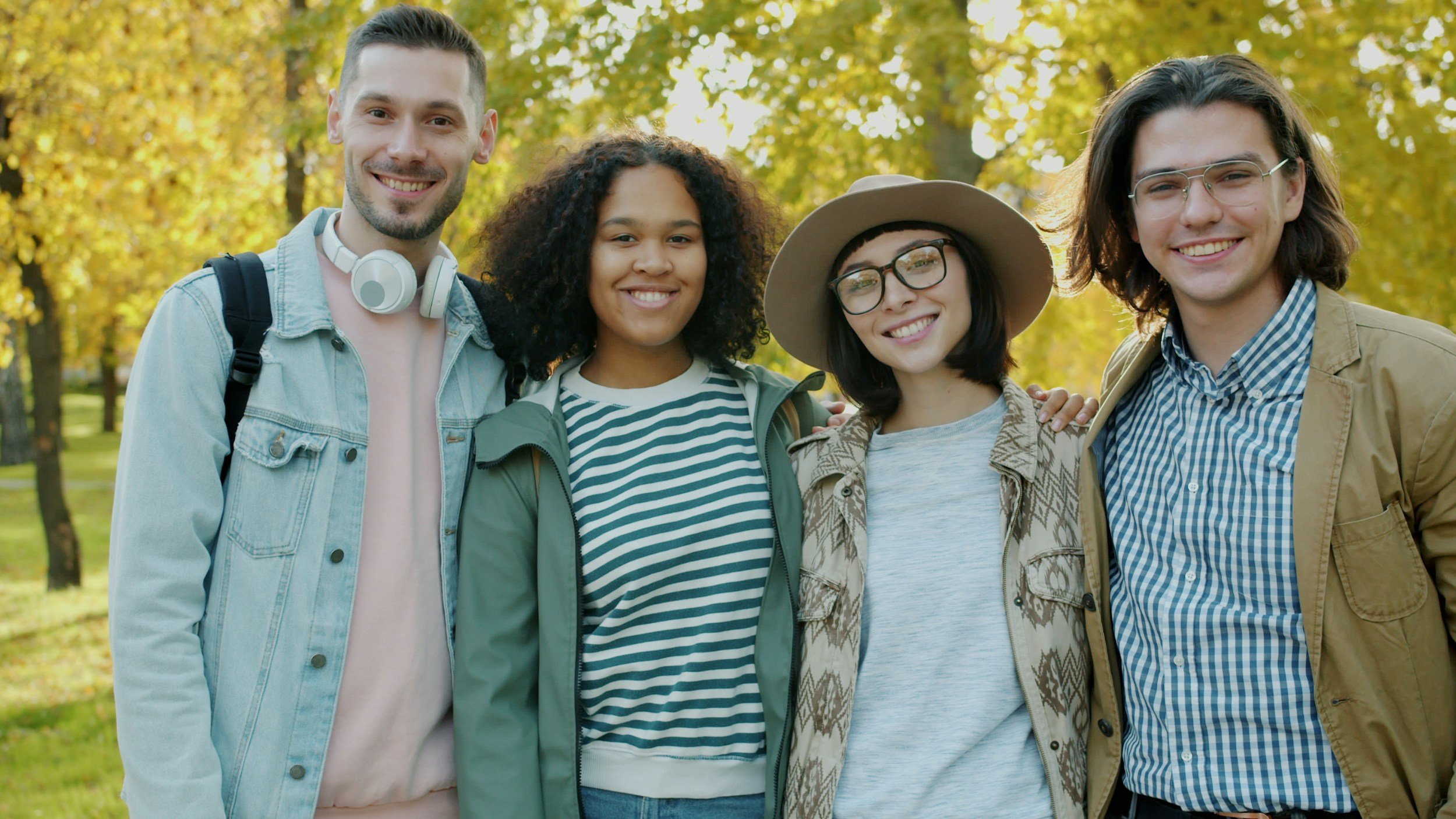 Four friends standing together outdoors in a park with autumn trees in the background, smiling at the camera.