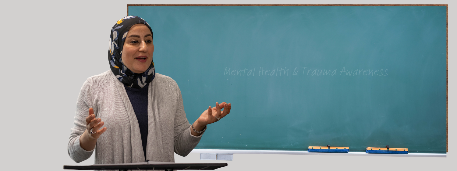 A woman wearing a headscarf and a beige cardigan speaking in front of a green chalkboard that reads 'Mental Health & Trauma Awareness'.