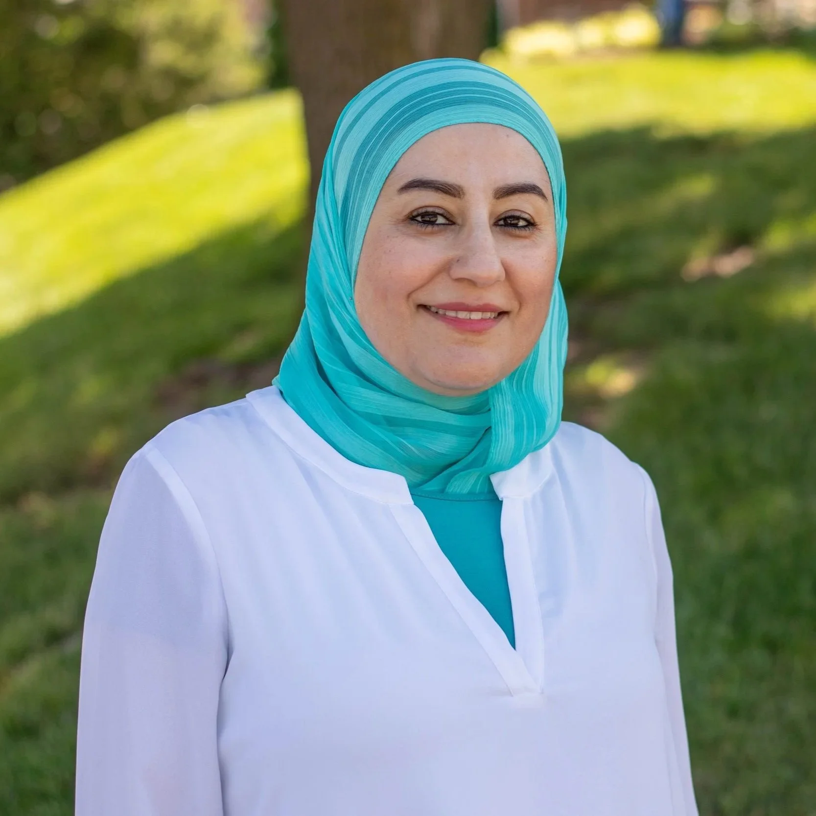 A woman in a blue hijab and white top standing outdoors with greenery and a tree in the background, smiling at the camera.