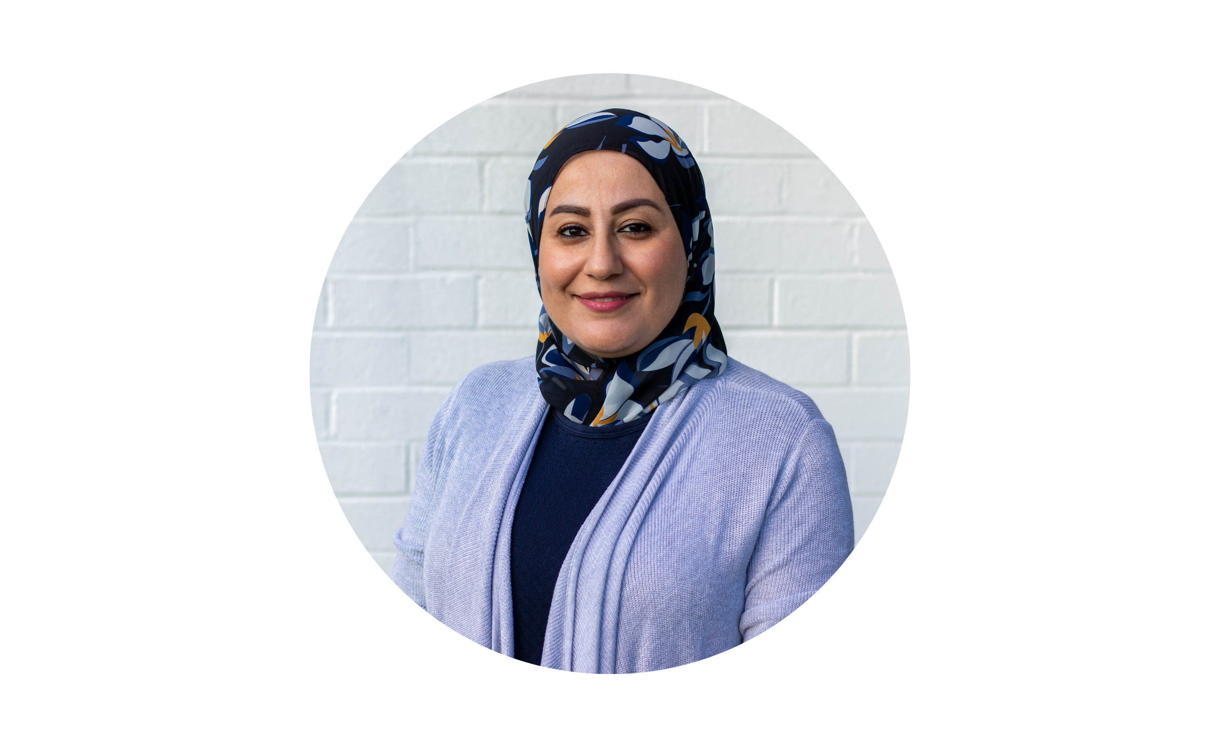 Rabab LMFT wearing a patterned headscarf and a light purple cardigan, standing against a white brick wall, smiling at the camera.