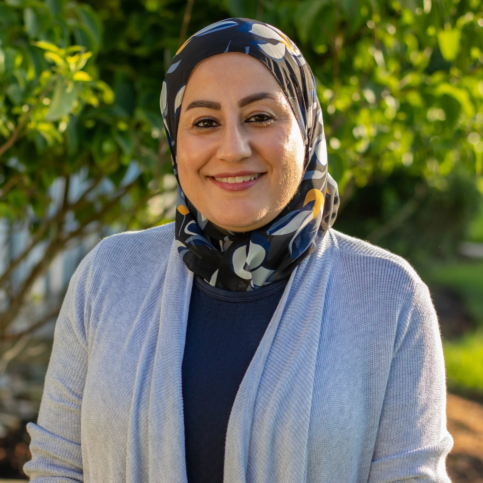 A woman smiling outdoors wearing a patterned headscarf and light-colored cardigan with greenery in the background.