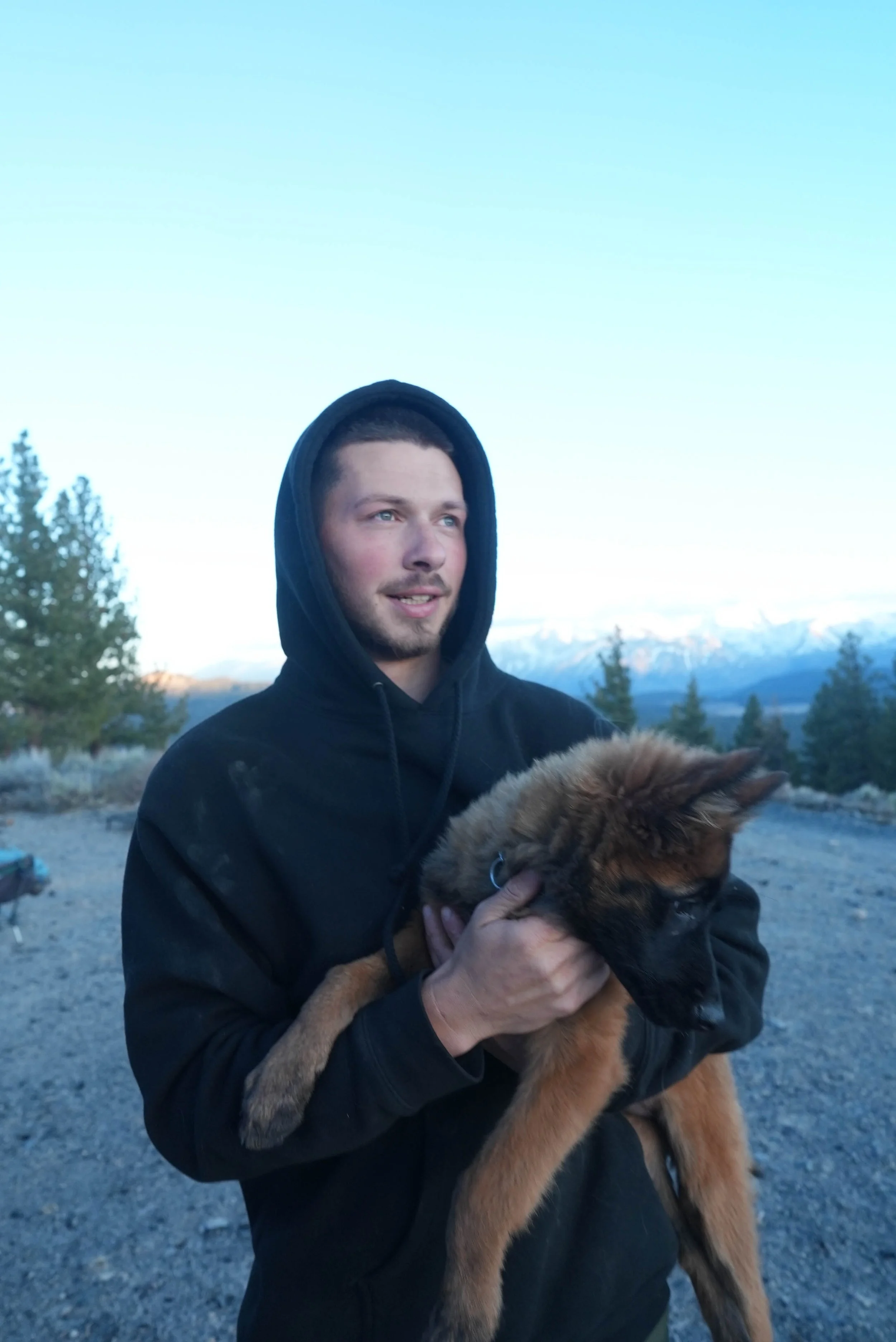 A man in a black hoodie holding a fluffy brown and black puppy outdoors with mountains and trees in the background.
