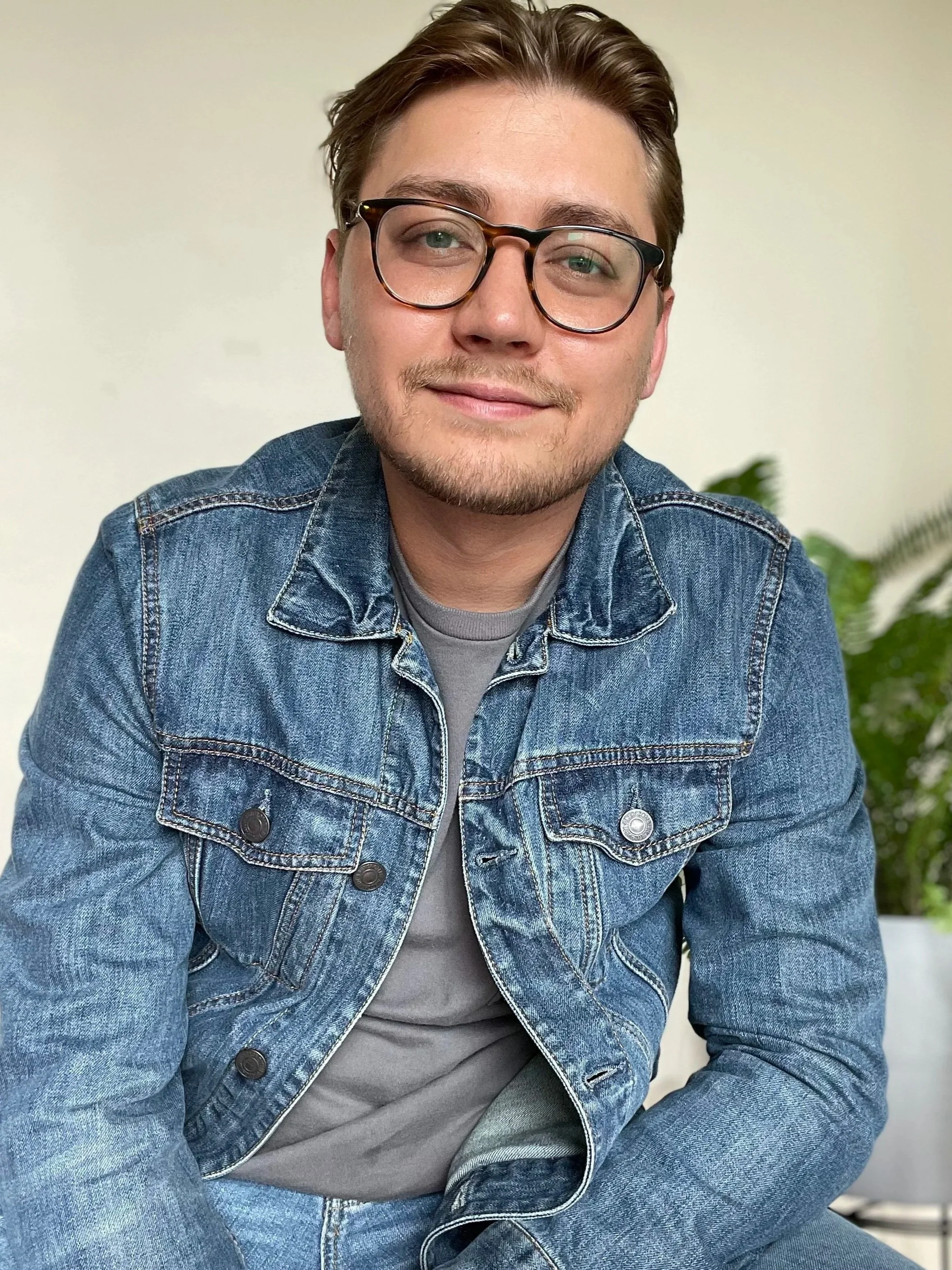 A young man with brown hair, glasses, and a light beard, smiling at the camera, wearing a blue denim jacket and a gray t-shirt, sitting indoors with a plain light-colored wall and some green plants in the background.