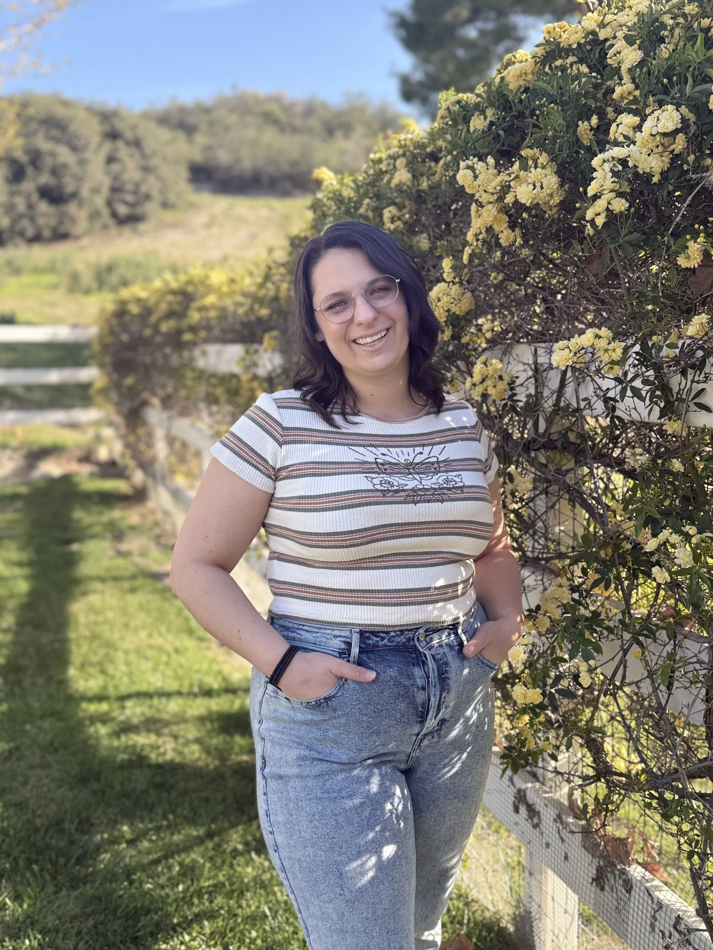 A young woman with dark hair and glasses standing outdoors next to a bush of yellow flowers, smiling. She is wearing a striped t-shirt and high-waisted jeans.