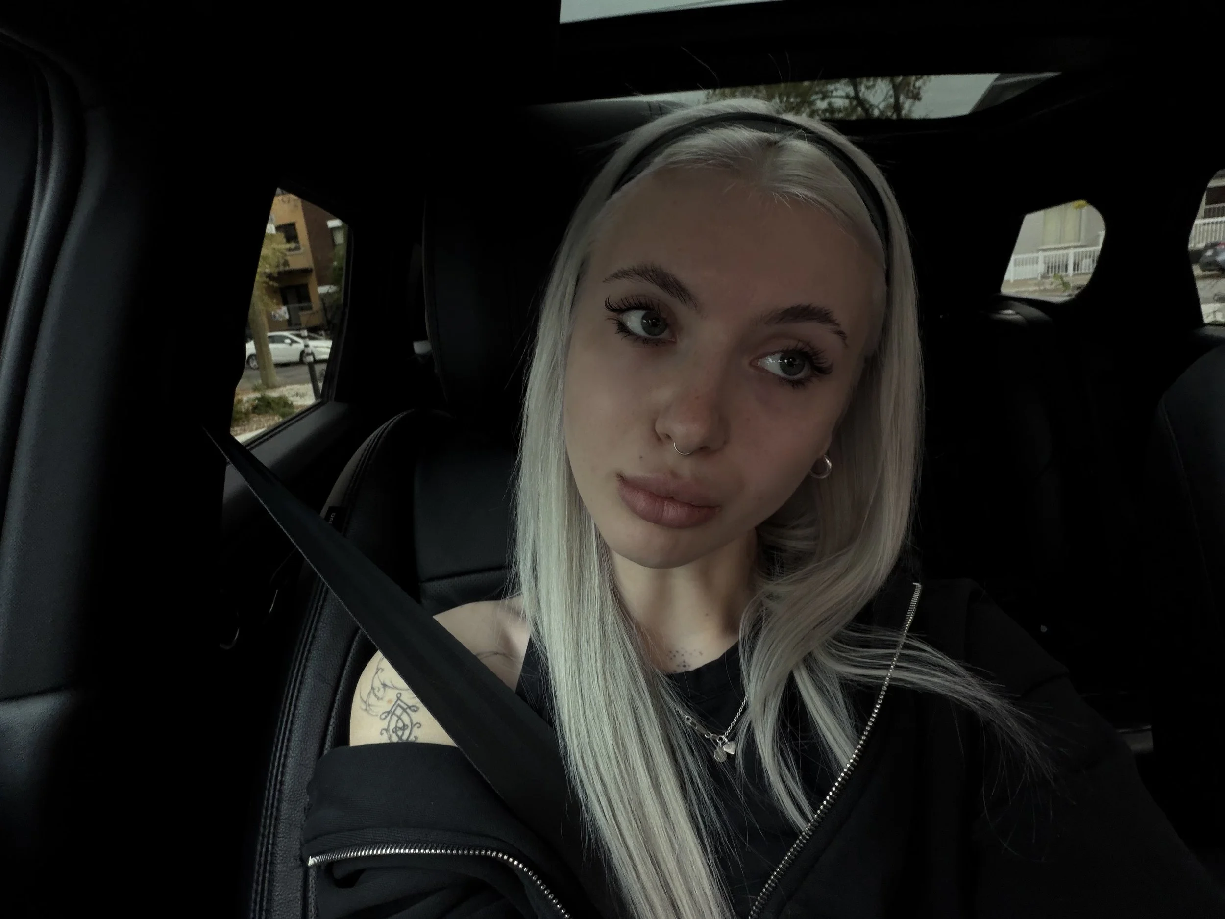 A young woman with long, platinum blonde hair and a septum piercing sitting in the backseat of a car, wearing a black top and a zip-up jacket.