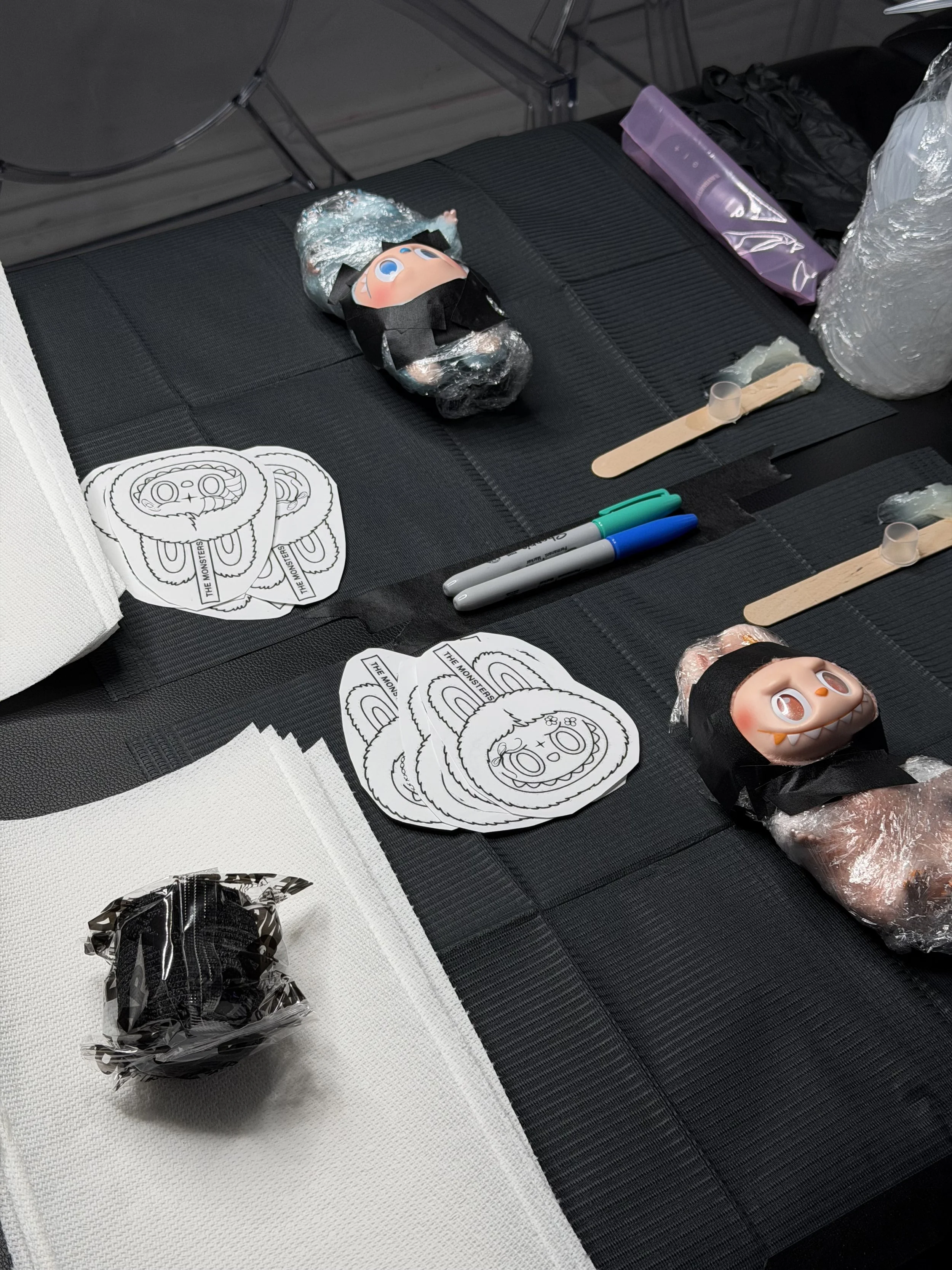Tattoo supplies on a black table , printed coloring pages, markers, wooden sticks, tape, and a sealed black wrapped item.