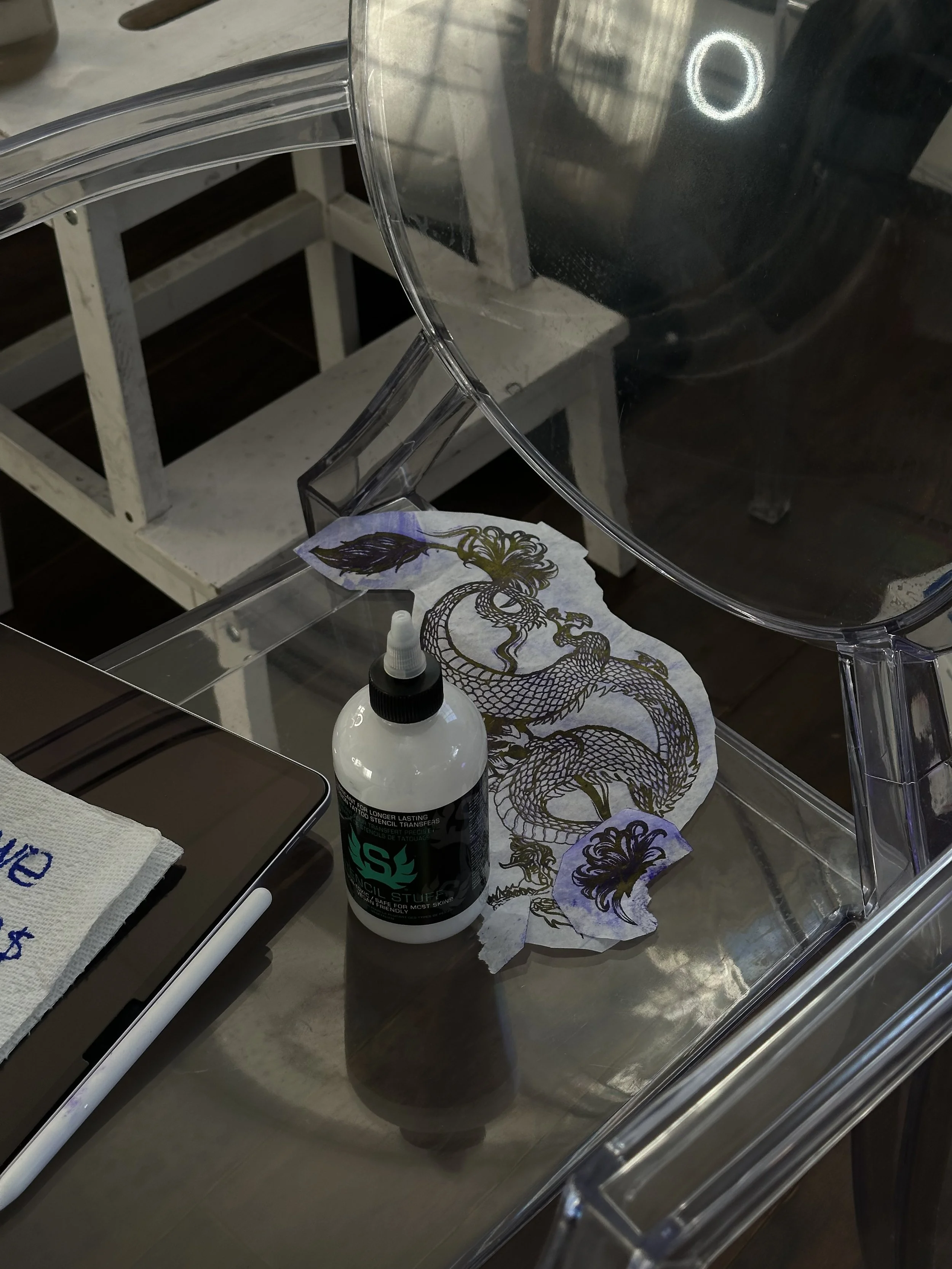 Tattoo stencil transfer paper with a cybercigilism dragon design, a white bottle of stencil transfer solution, and a tablet on a clear acrylic surface.