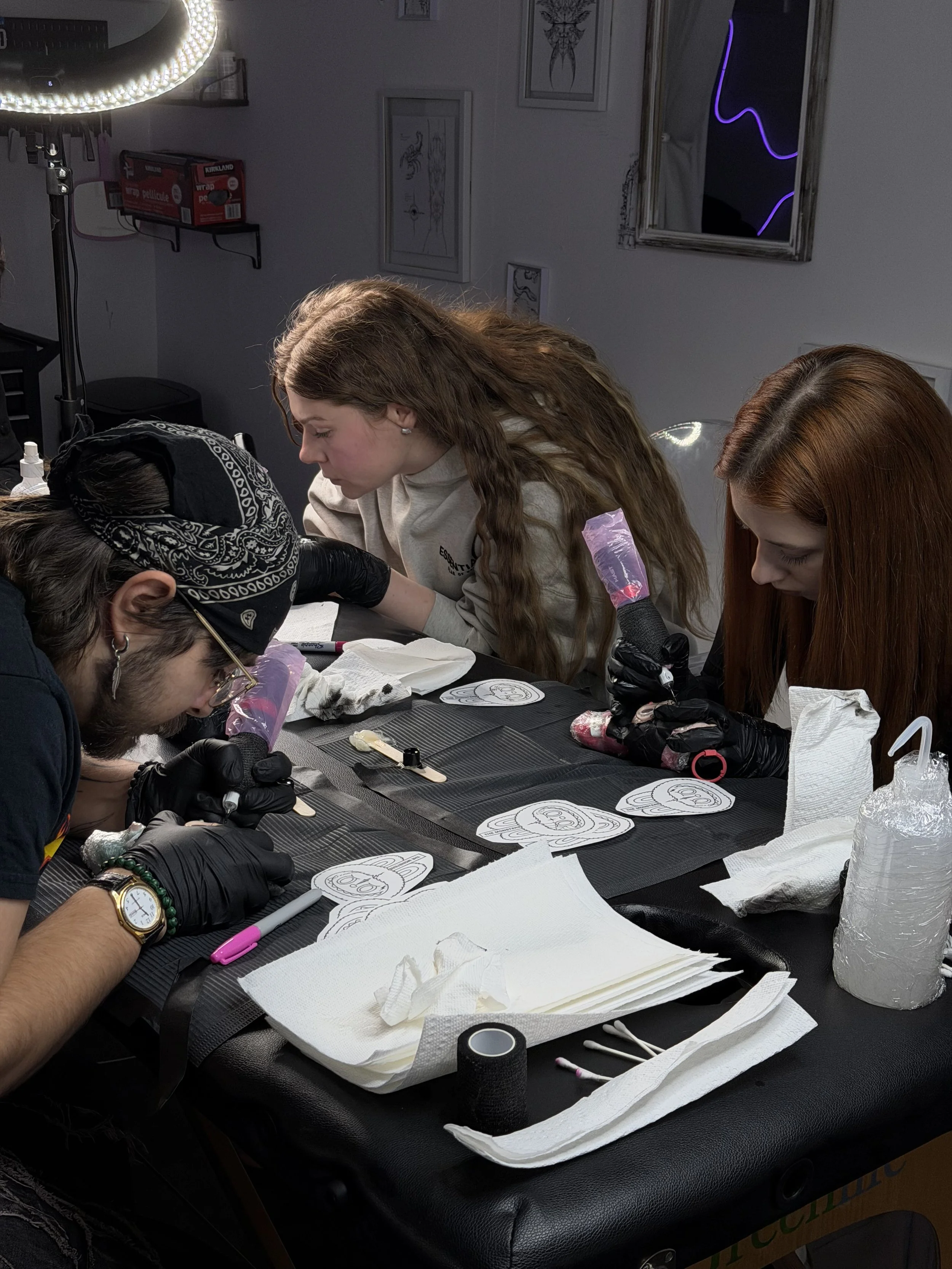 People engaged in tattooing practice on fake skin, surrounded by tattoo supplies in a studio.