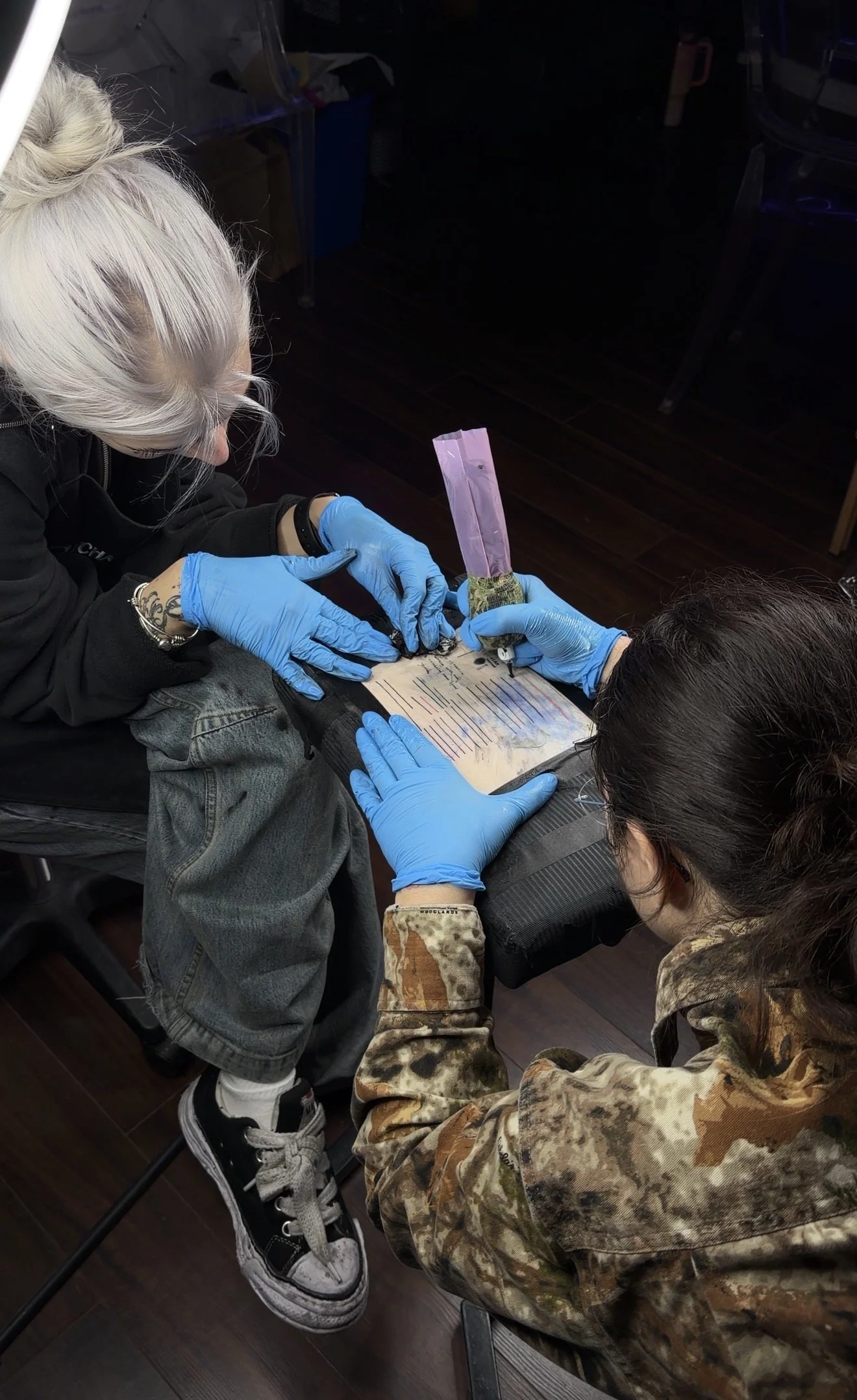 Two people with blue gloves tattooing fake skin, seated in a dark room.