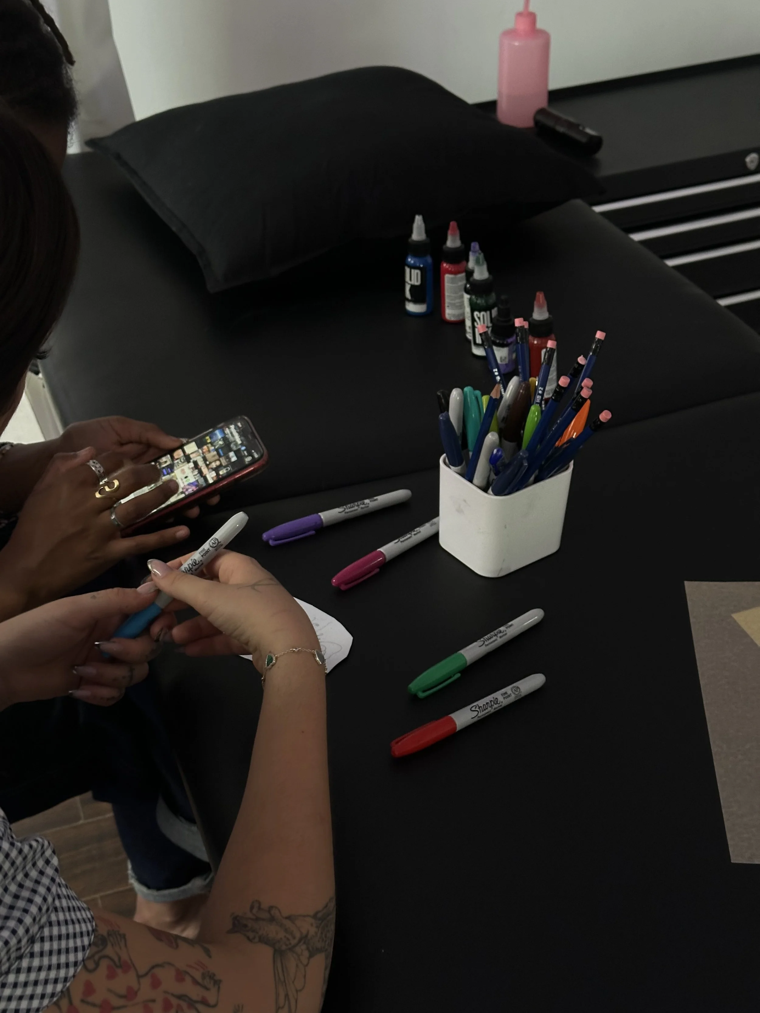 A black table with a white container holding various colored pens, some colored markers, and a few bottles of ink. Two people are sitting at the table, one using a smartphone and the other using a marker, with tattoos visible on their arms. There are additional markers on the table and a pink spray bottle in the background.
