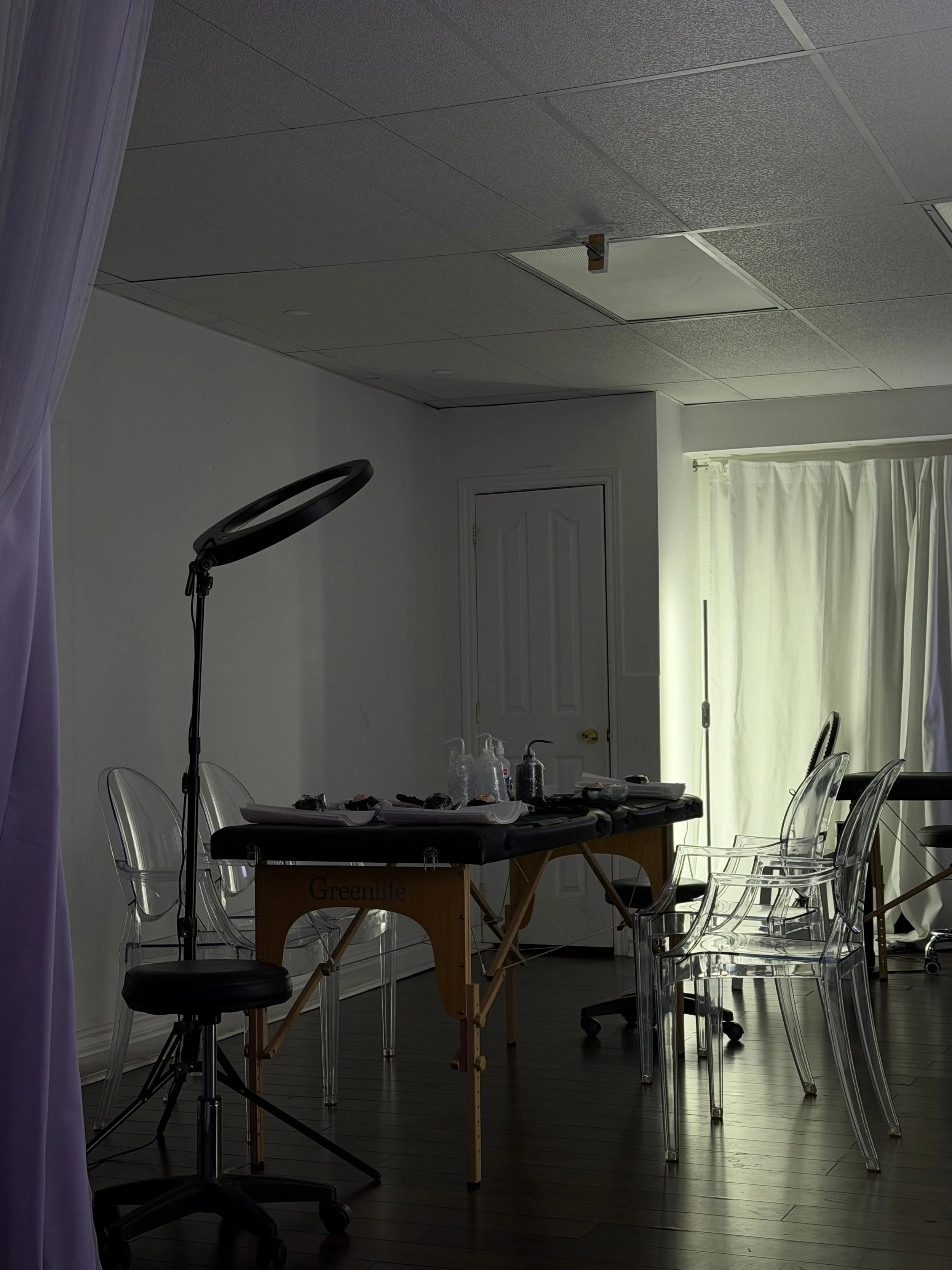 Tattoo studio with clear plastic chairs, a work table with supplies, and a ring light on a stand, with a closed door and curtains in the background.