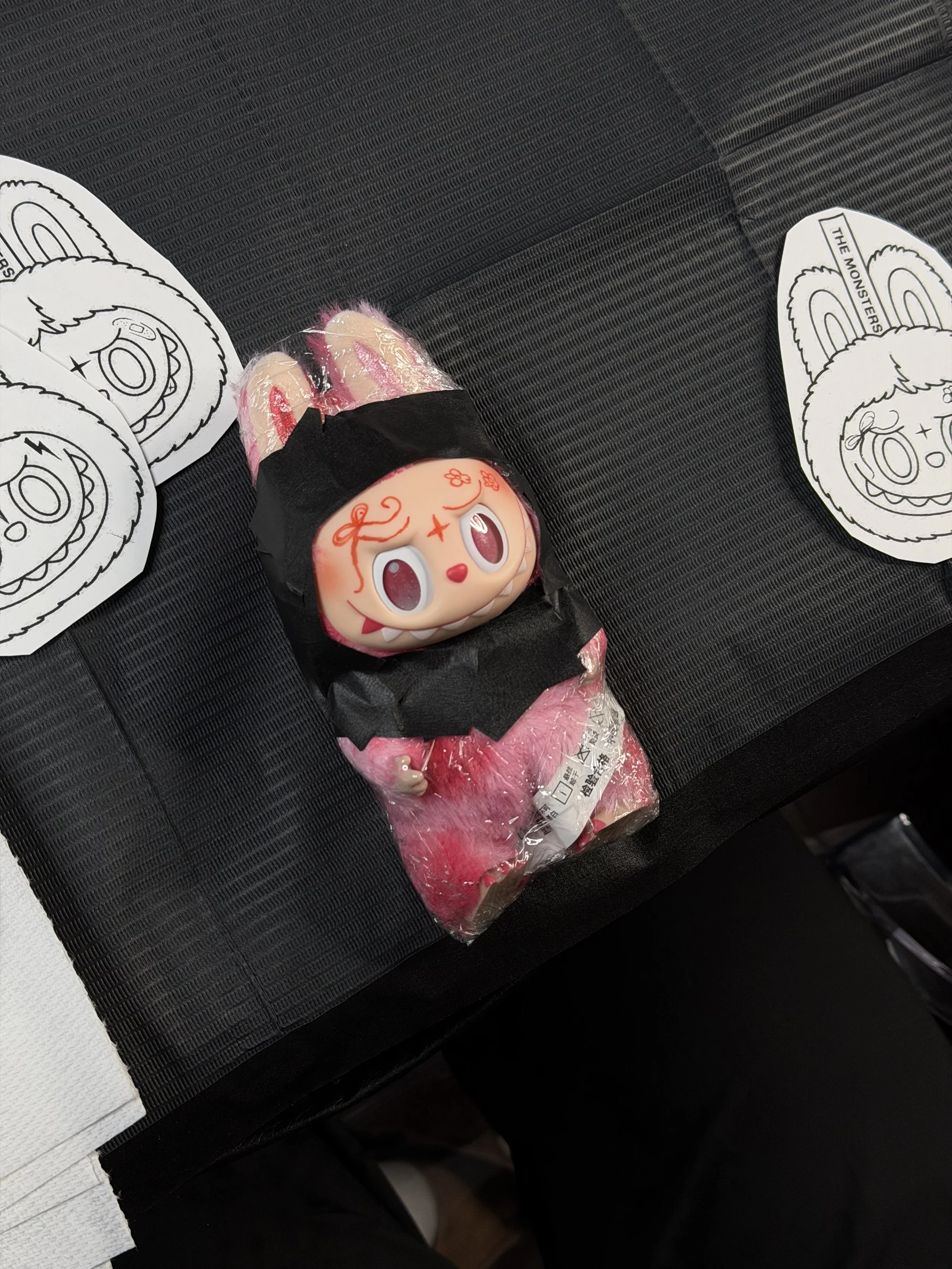 Labubu, wrapped in plastic and secured with black tape, placed on a black surface with paper cutouts of a smiling face with a headband and the text "THE MONSTERS."