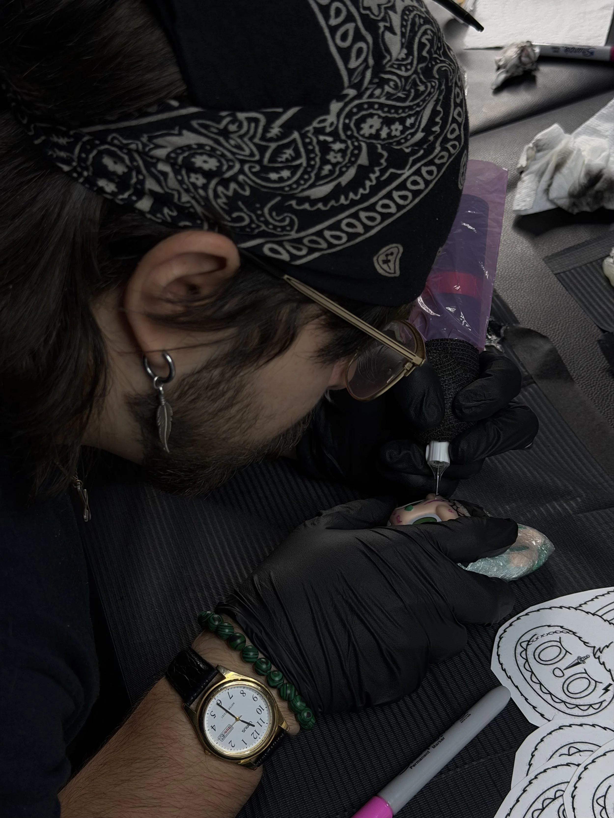 A person with a bandana, earrings, and glasses is tattooing a colorful cartoon-like face on a Labubu or artificial skin, wearing black gloves, with tattoo supplies and drawings nearby.