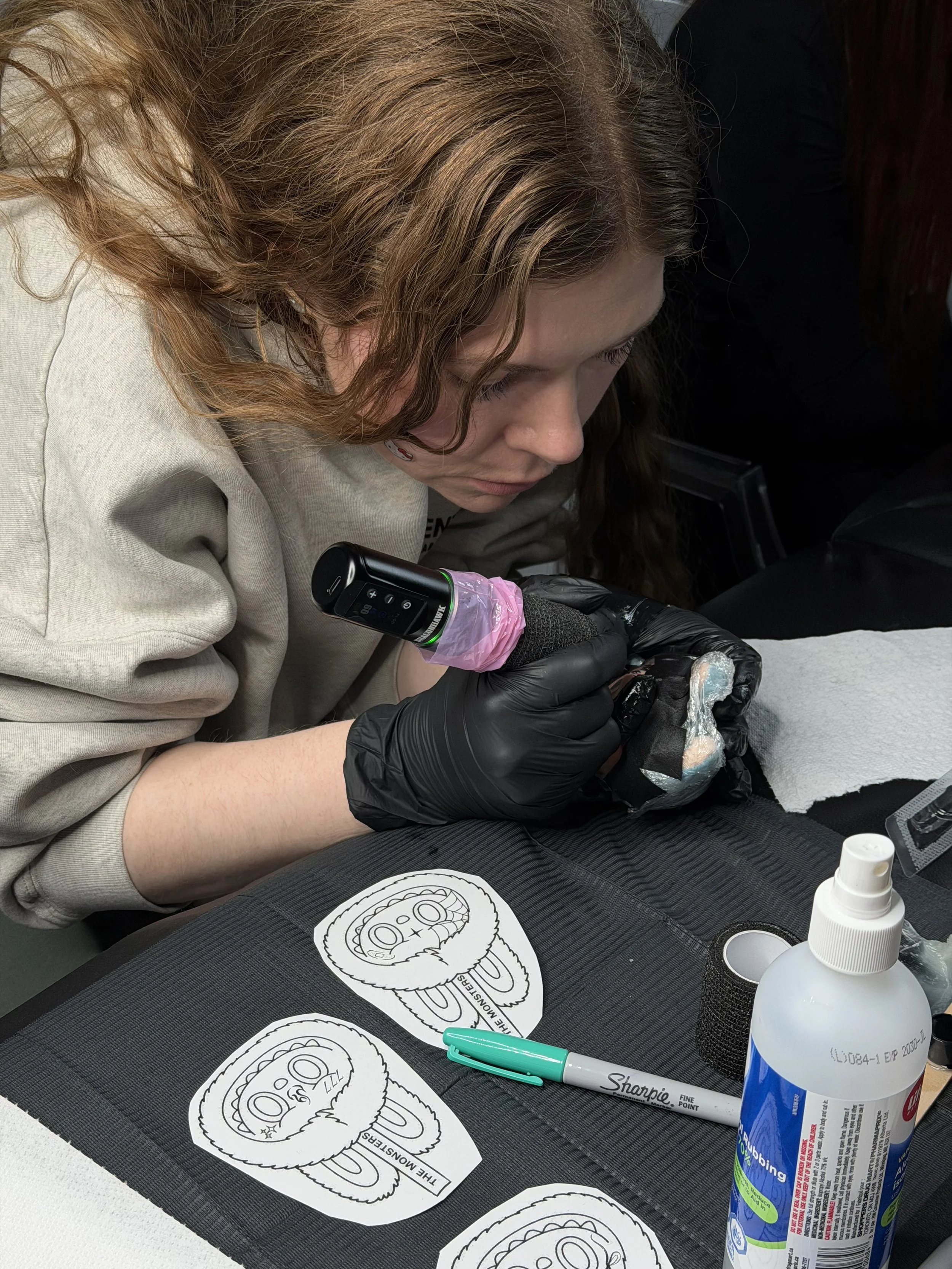 A woman with curly hair and black gloves is tattooing a design on aLabubu. There are tattoo stencil sheets with a cartoonish face of Labubu on the table, along with a Sharpie marker and a bottle of disinfectant spray.
