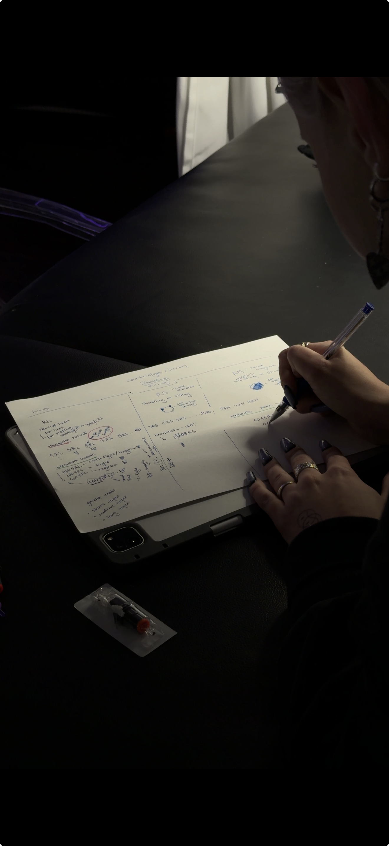 A person with dark nails and rings writing notes on a paper pad with handwritten notes and diagrams, with a tablet underneath, on a dark surface. 