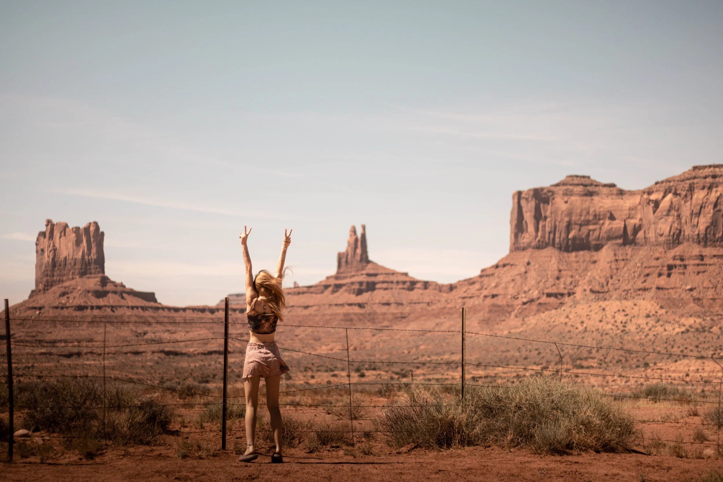 A woman standing in front of a desert landscape with rock formations, raising her arms in a V shape as the sun shines.