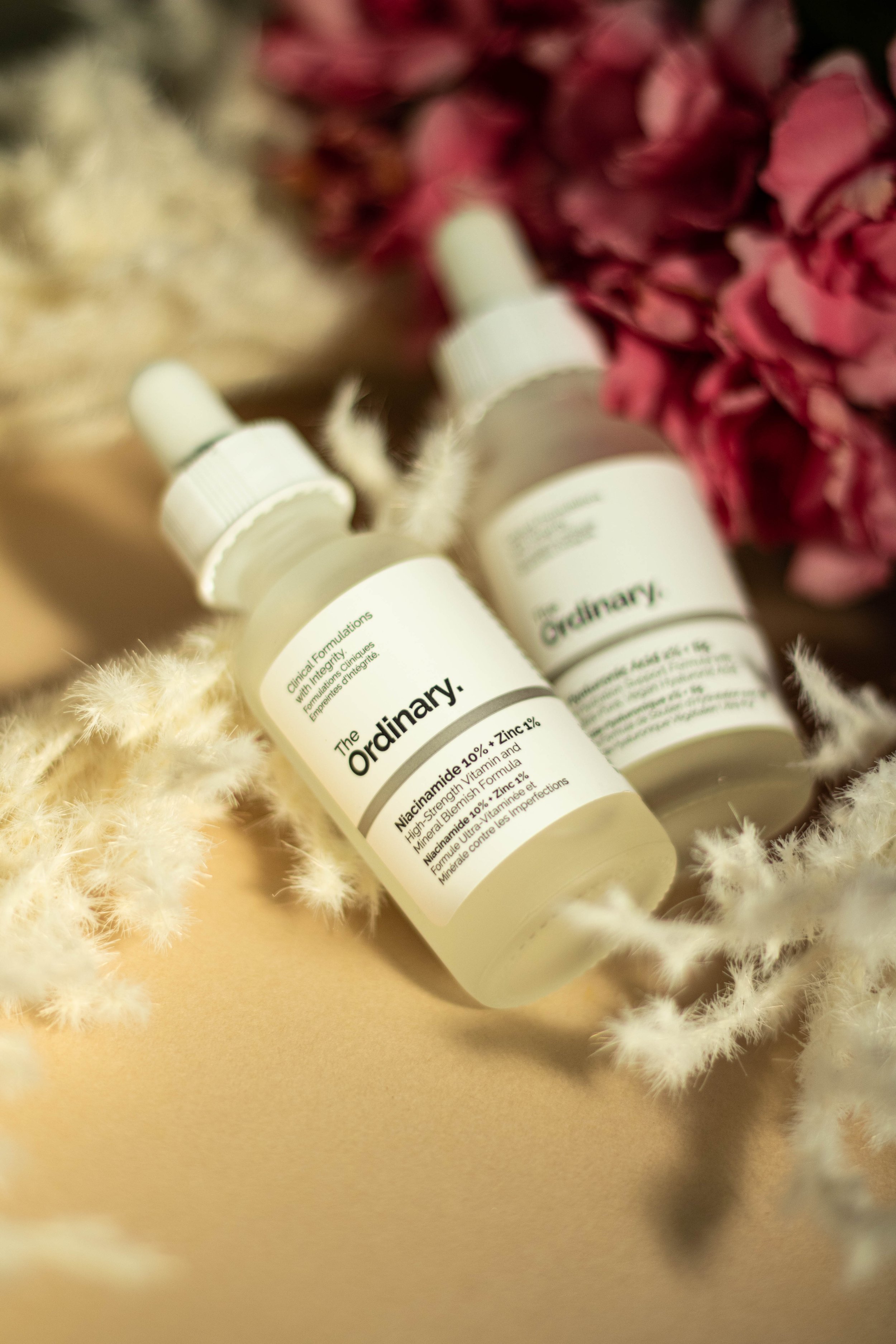 Close-up of two skincare bottles from The Ordinary placed on a beige surface, surrounded by soft white fluffy material and pink flowers in the background.