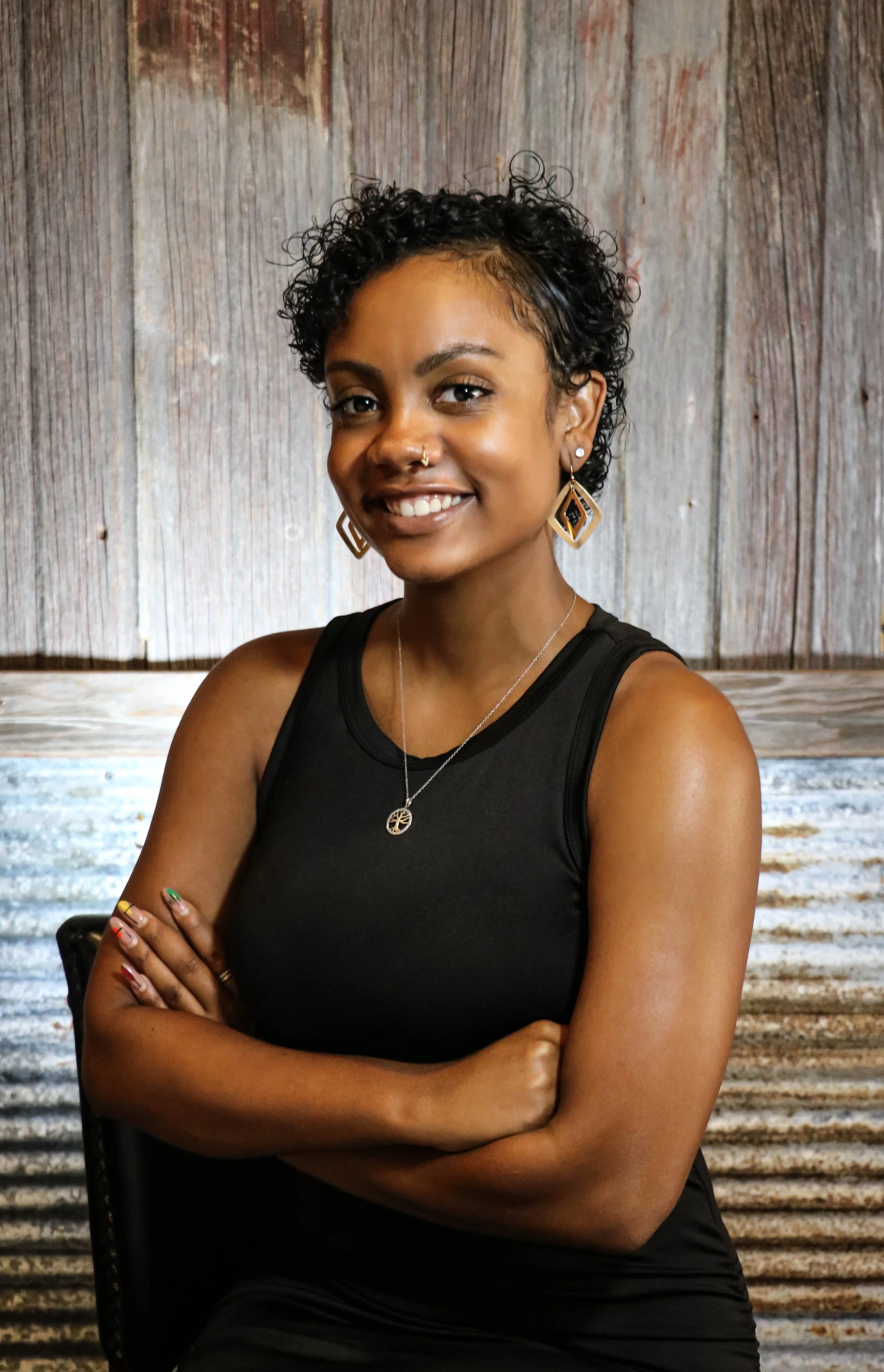 A young woman with short curly hair, wearing a black sleeveless top, earrings, a necklace, and colorful nail polish, sitting with her arms crossed in front of a wooden background.