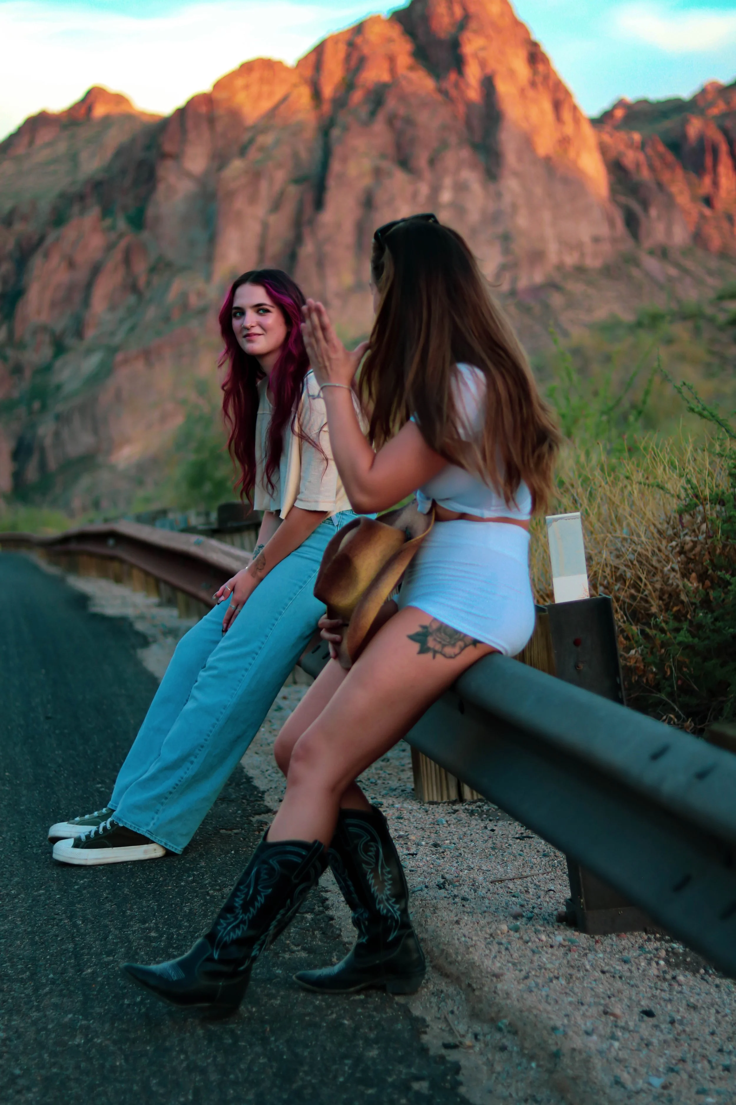 Two young women sitting on a roadside guardrail with red rock mountains in the background, during sunset. One woman has long pink hair, and the other has long brown hair and tattoos on her thigh. They are dressed casually, with one wearing cowboy boots and the other wearing sneakers.