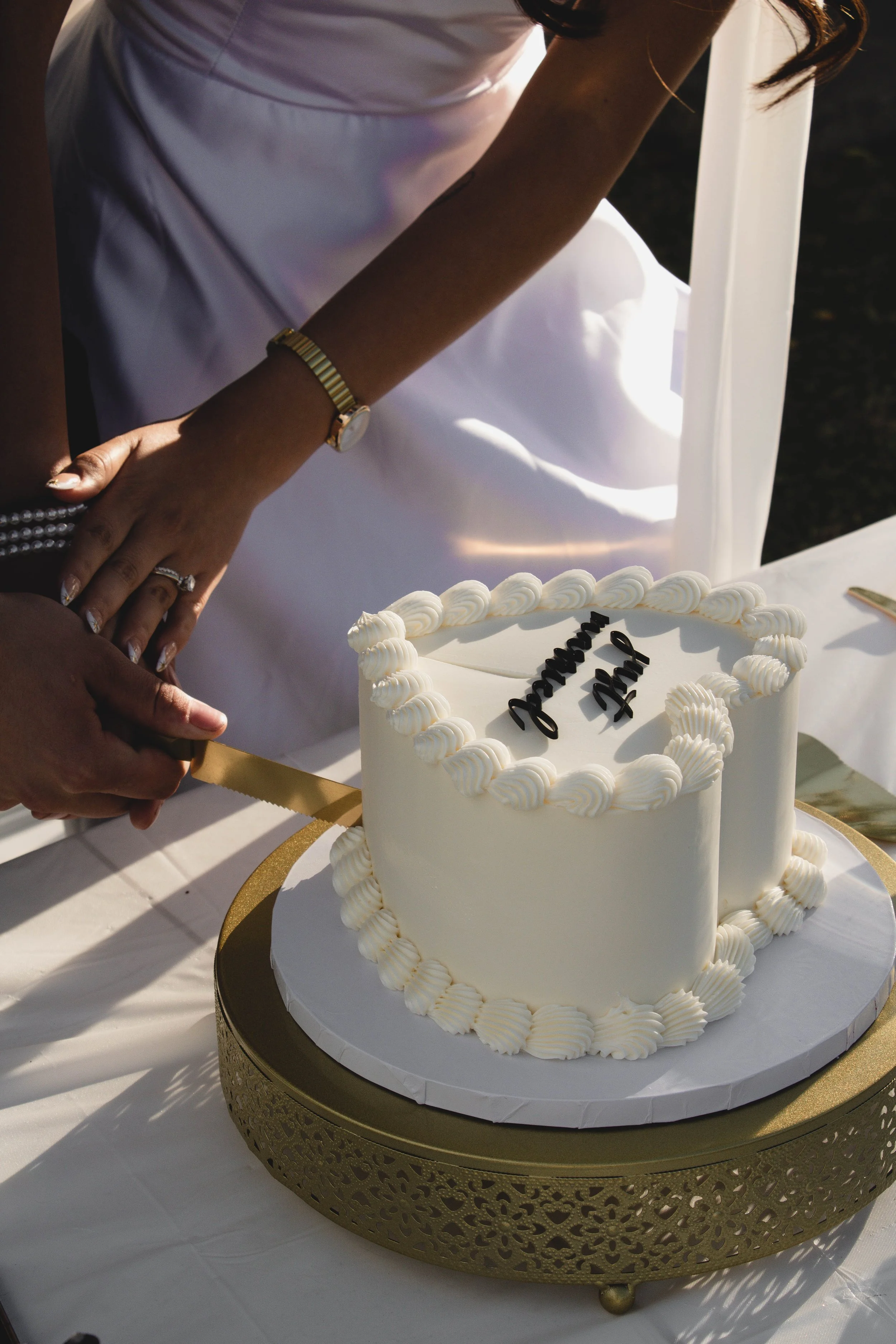 Close-up of a wedding cake with white icing and decorative piping, placed on a gold-colored decorative stand, with a woman in white dress visible in the background.