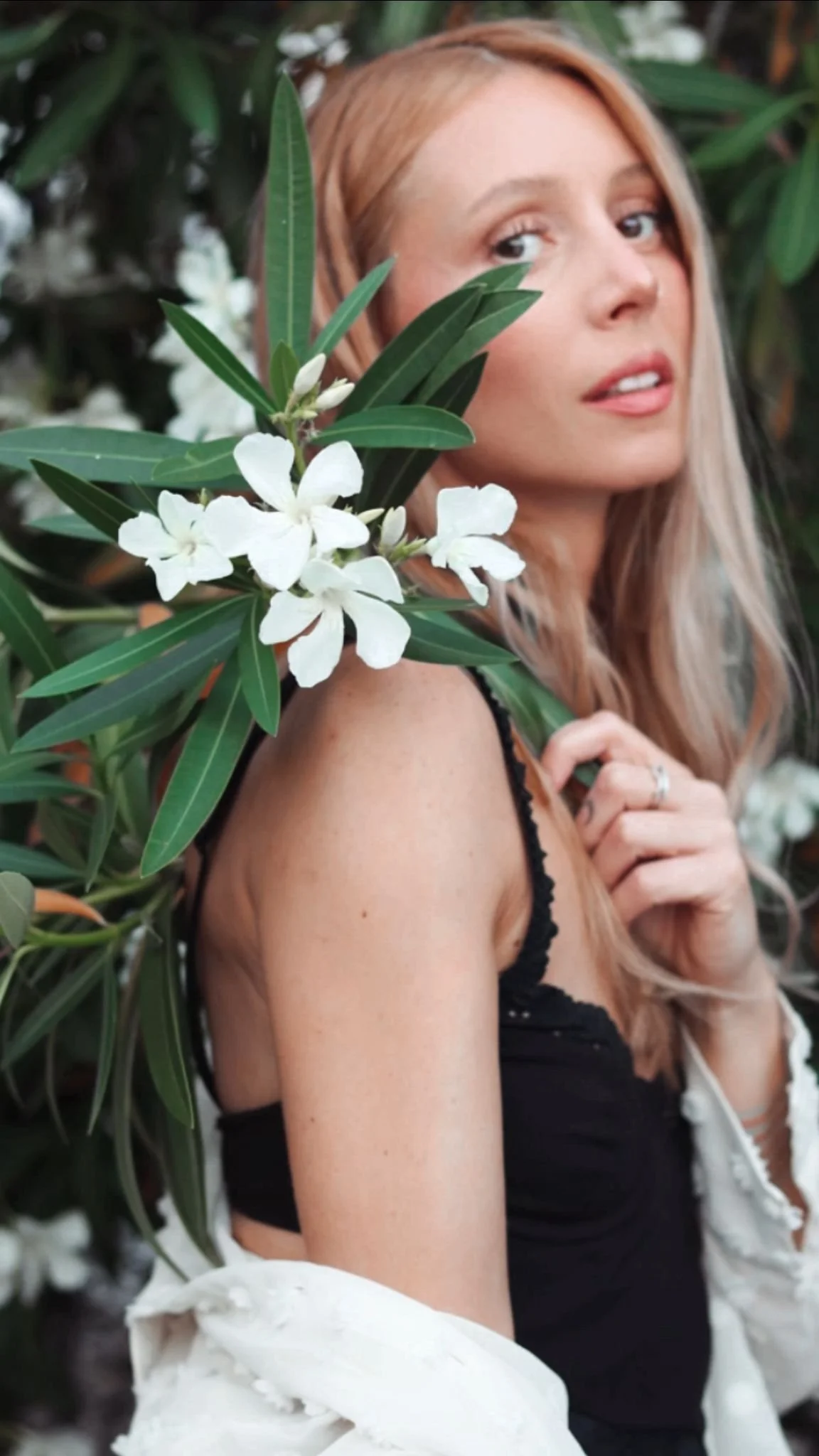 A woman with reddish-blonde hair partially obscured by green leaves and white flowers, wearing a black top and a white jacket, looking into the camera with a gentle expression.