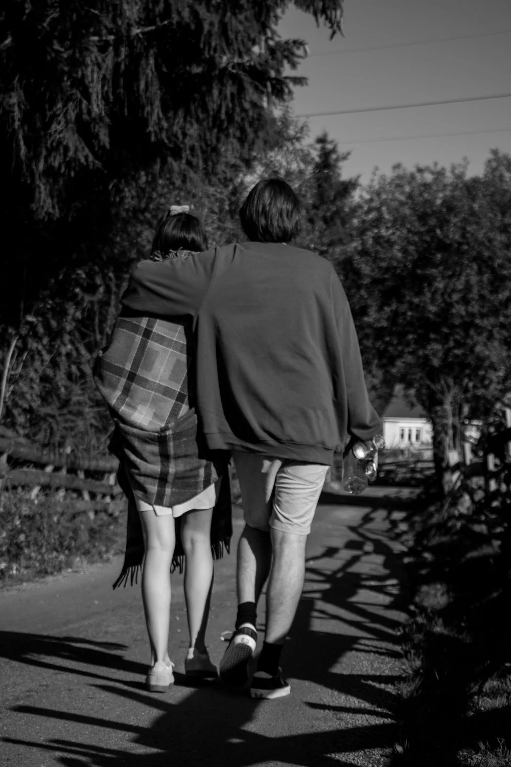 Two people walking on a sidewalk, seen from the back, with trees lining the path. One person has an arm around the other.