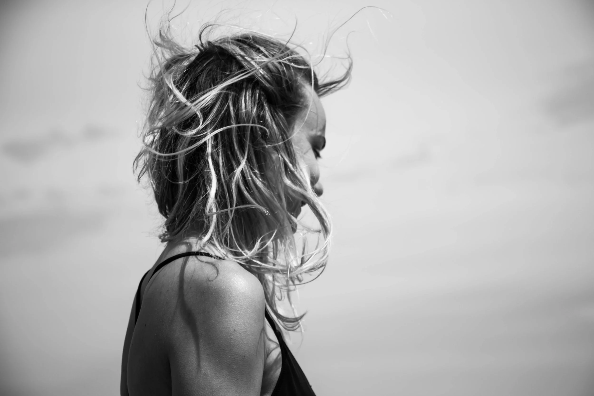 Black and white photo of a woman with wavy hair looking to the side, wearing a sleeveless top, against a plain sky background.