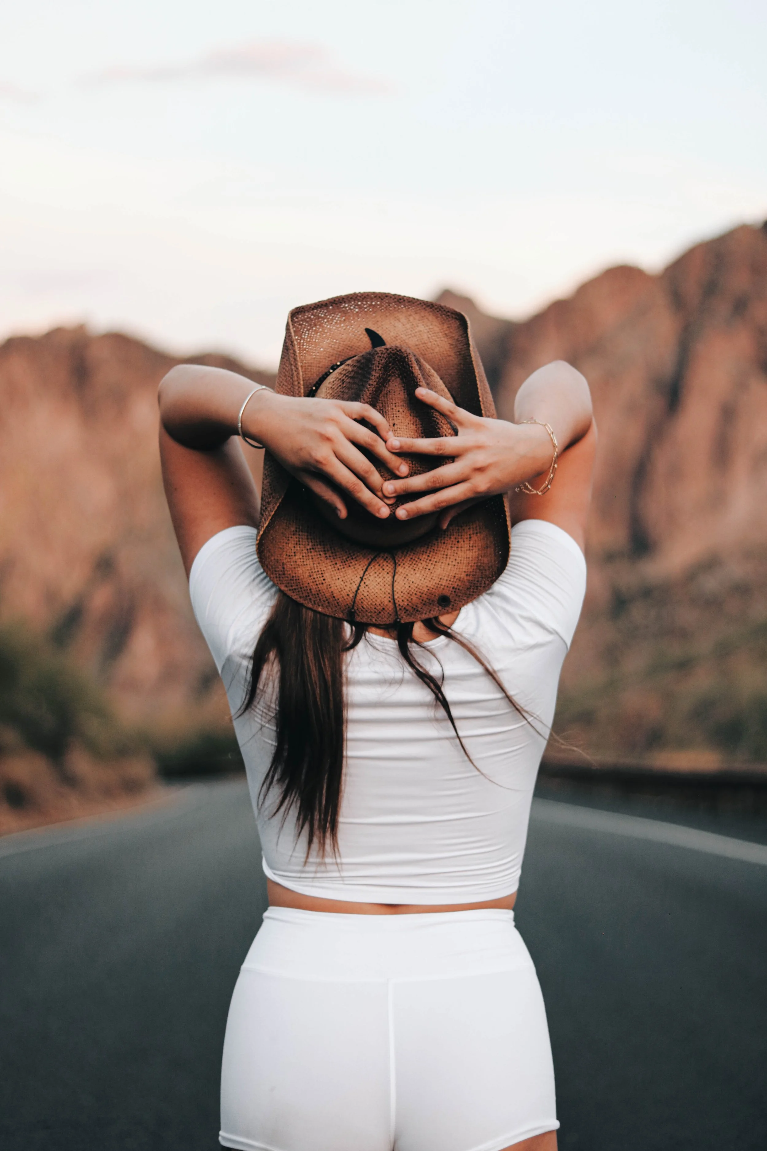 Person standing on a road in front of red rock formations, wearing a white crop top, white shorts, a brown hat, and holding the hat with both hands, facing away from the camera.