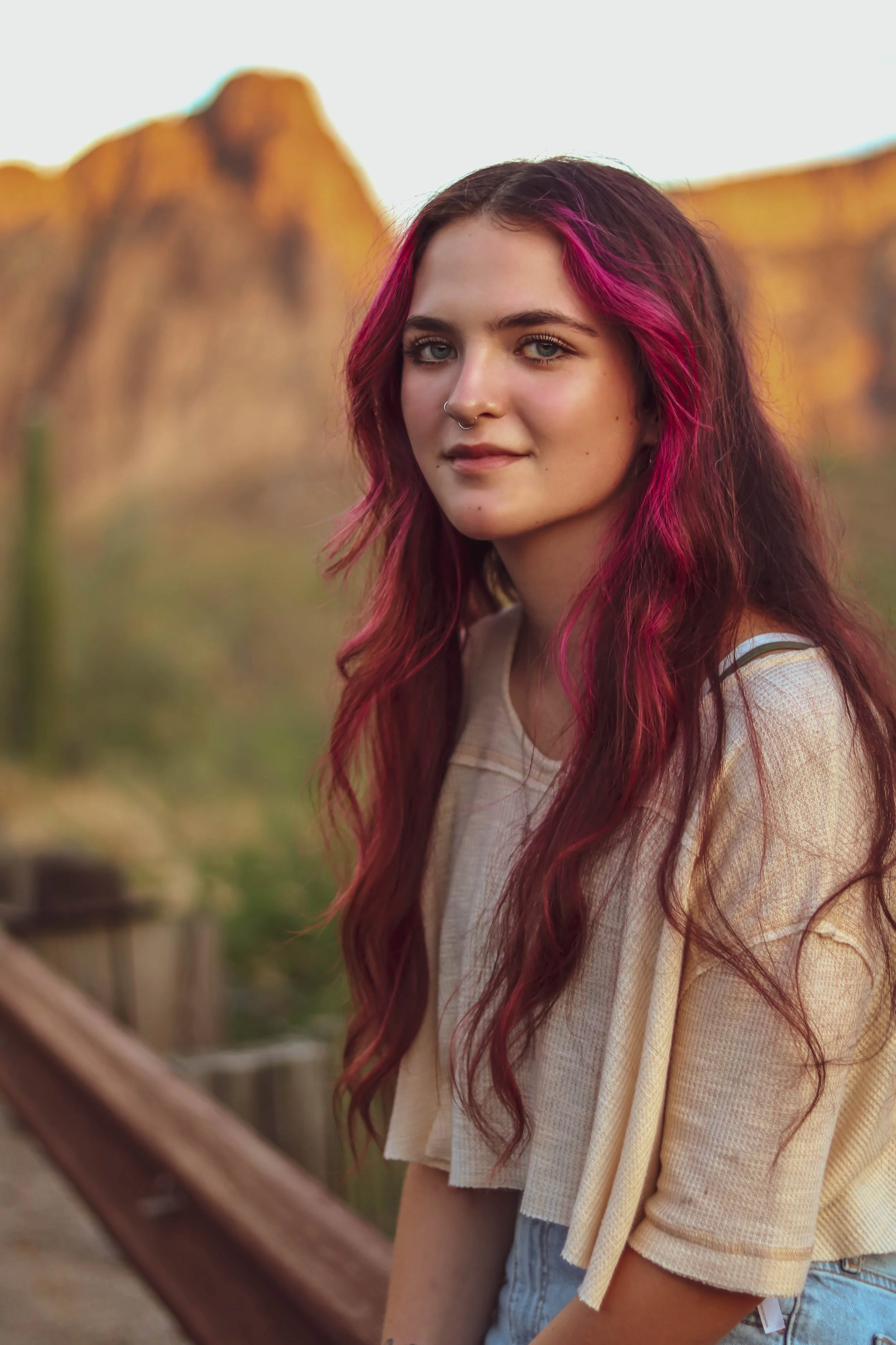A young woman with long dark hair and pink highlights, wearing a nose piercing and a beige top, sitting outdoors near a wooden fence with a mountainous landscape in the background during daylight.