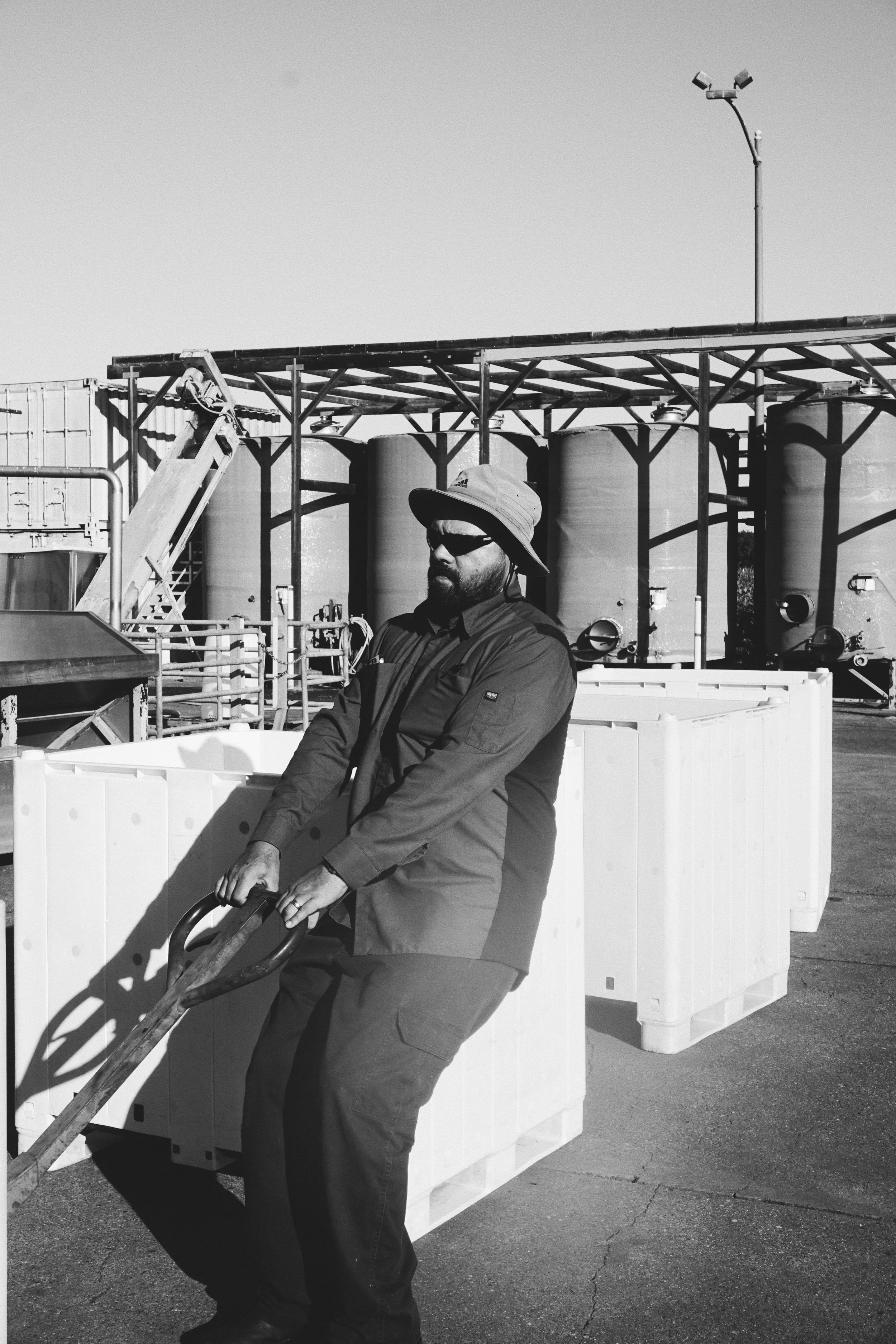 A man wearing a wide-brimmed hat, sunglasses, and outdoor work clothing, standing outdoors with large storage tanks and industrial equipment in the background.