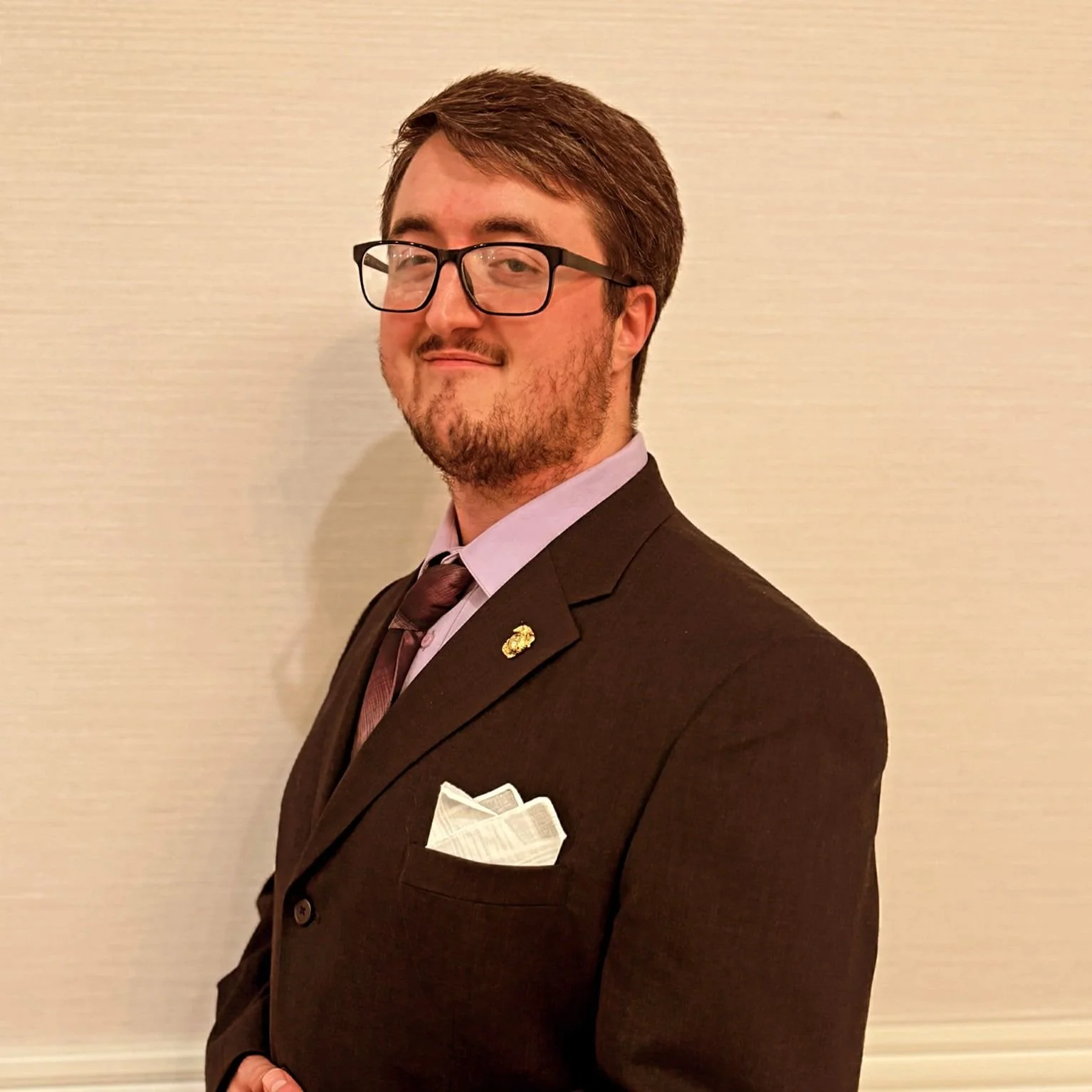 A young man with glasses, a beard, and brown hair, dressed in a dark brown suit with a light purple shirt, a matching tie, a white pocket square, and a small lapel pin, standing against a plain beige wall.
