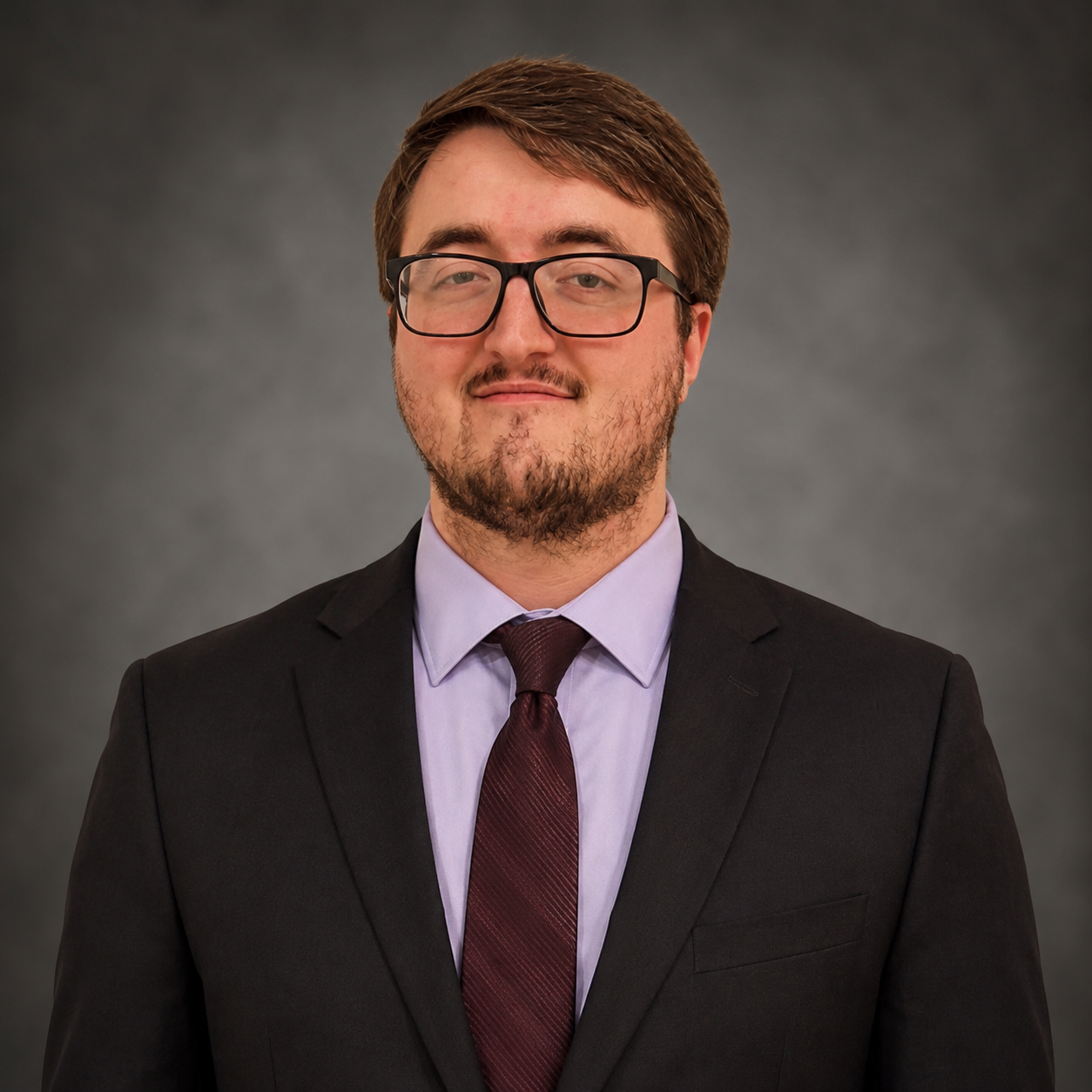 A young man with glasses, a beard, and brown hair, dressed in a dark brown suit with a light purple shirt, a matching tie, a white pocket square, and a small lapel pin, standing against a plain beige wall.