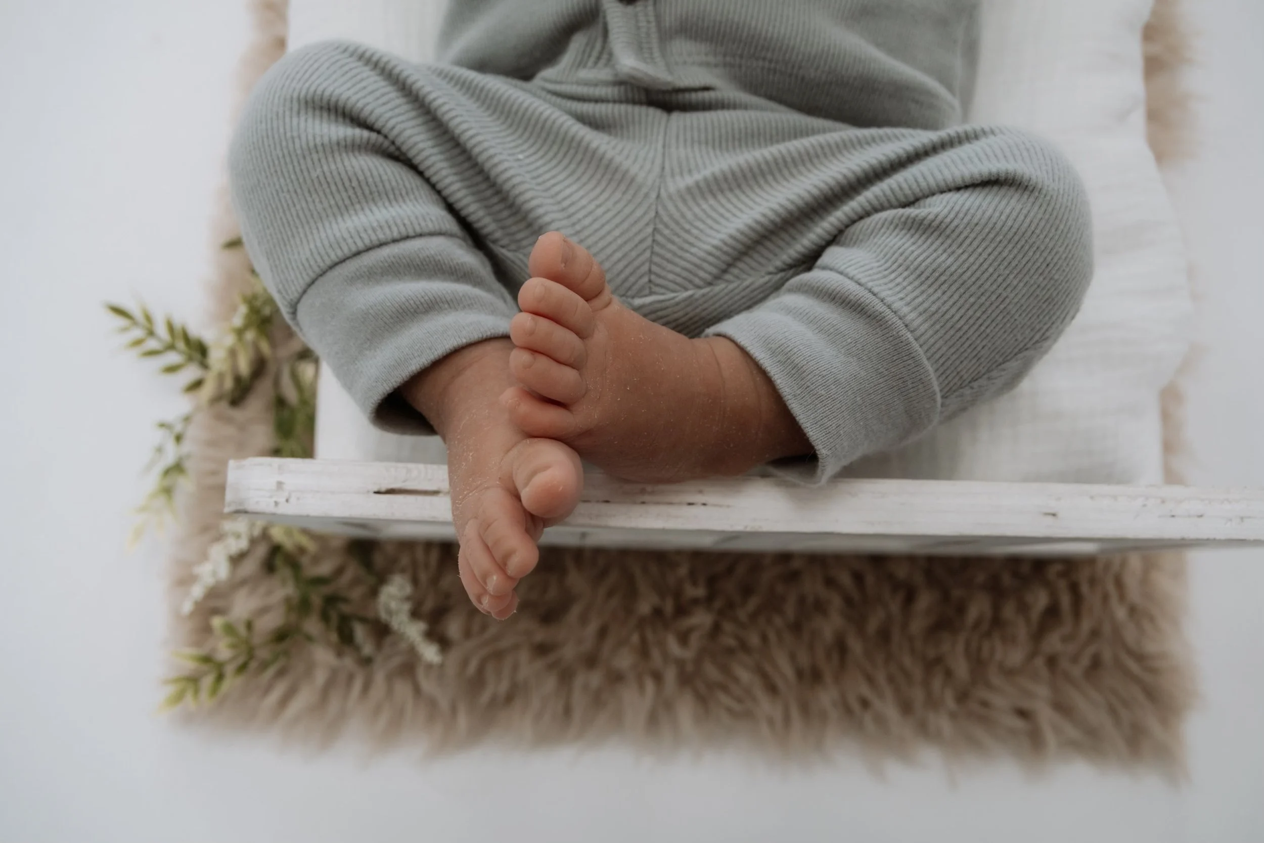Close-up of a baby's feet with one foot crossed over the other, placed on a white wooden surface, with a furry rug and decorative greenery underneath.