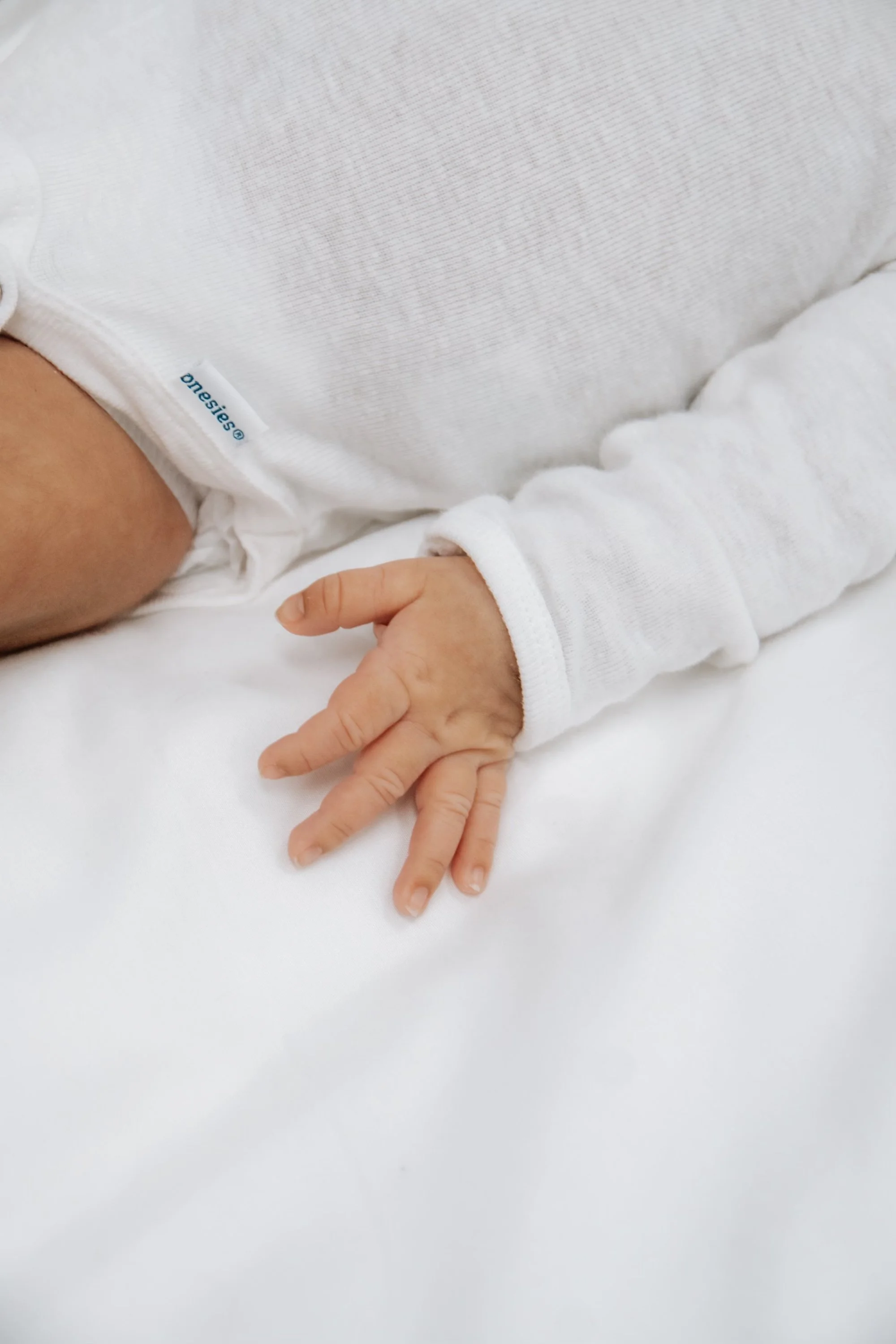 Close-up of a baby's hand resting on a white surface, with a white long-sleeve shirt.