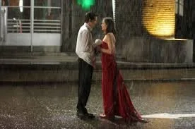 A man and woman standing under rain on a city street, facing each other, with the woman wearing a long red dress and the man in a white shirt and dark pants.