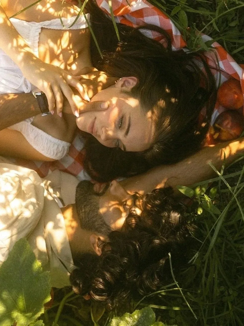 Two women lying on the grass and blanket, surrounded by plants and peaches, smiling and resting in sunlight with shadows of leaves on their faces.