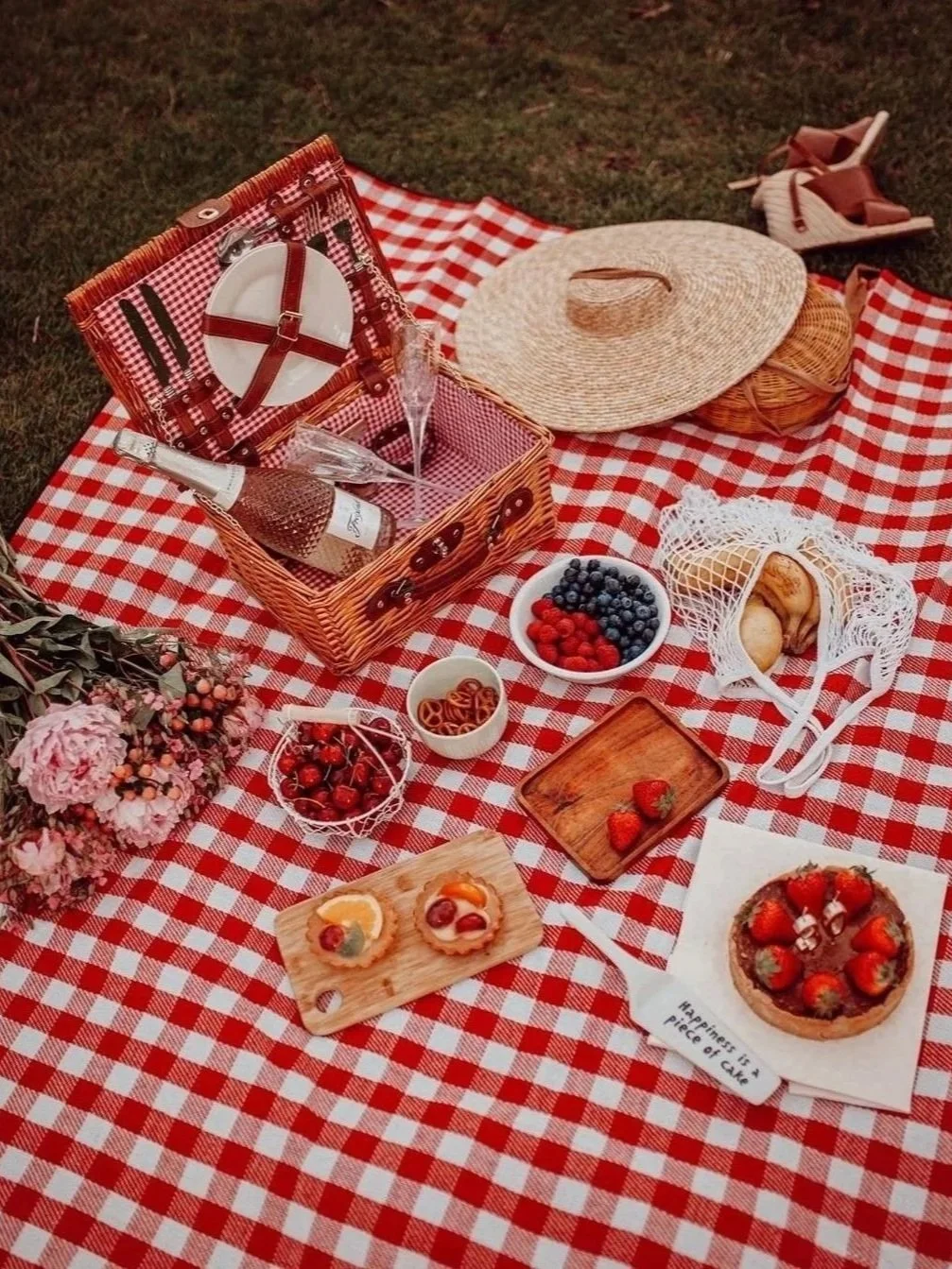 A picnic setup with a red and white checkered blanket, a wicker basket, bowls of berries and cookies, a cake with strawberries, a floral bouquet, a straw hat, and a pair of sandals.