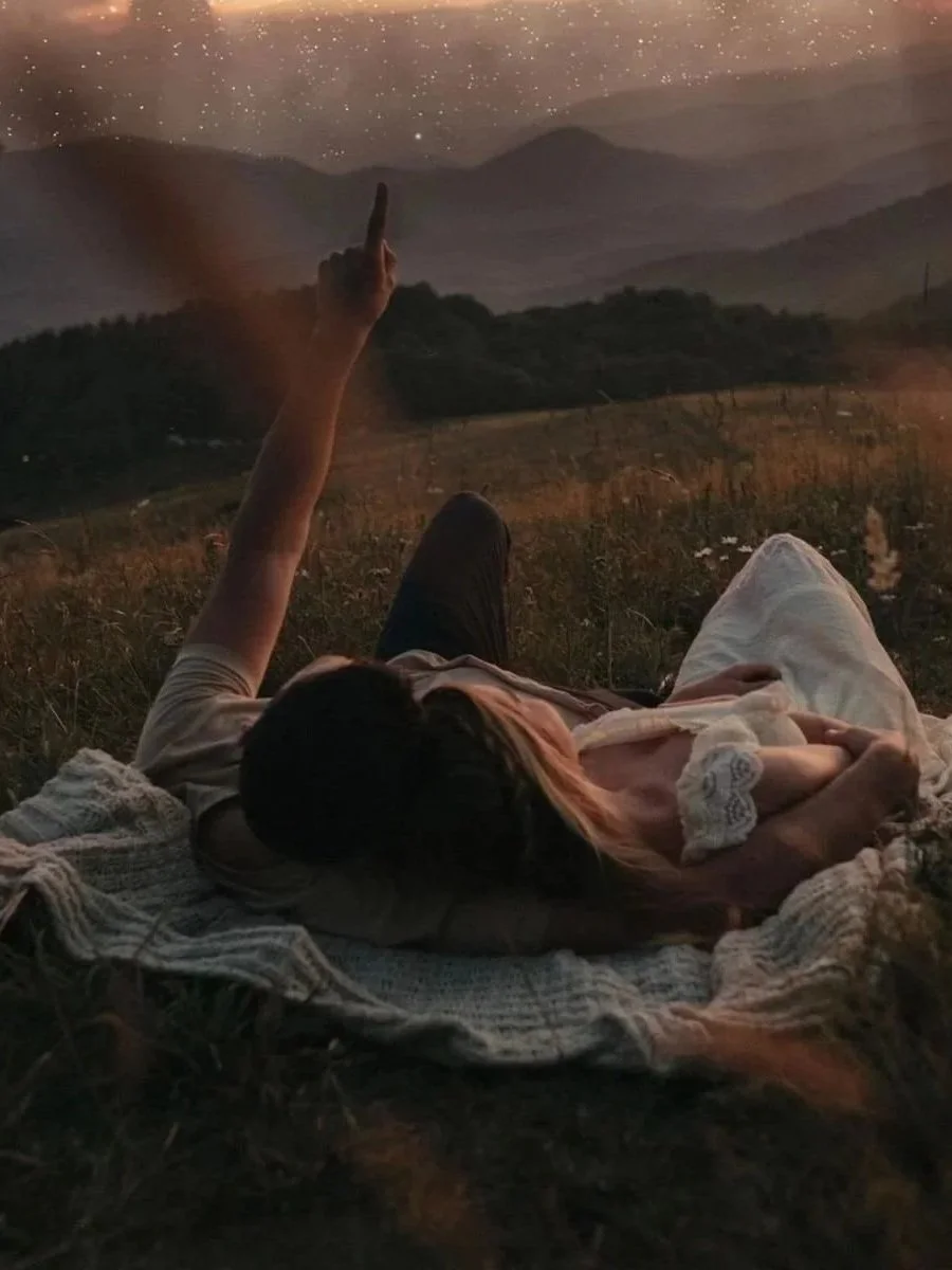 A couple lying on a blanket in a quiet grassy field, pointing toward a star-filled night sky, immersed in a peaceful moment of wonder.