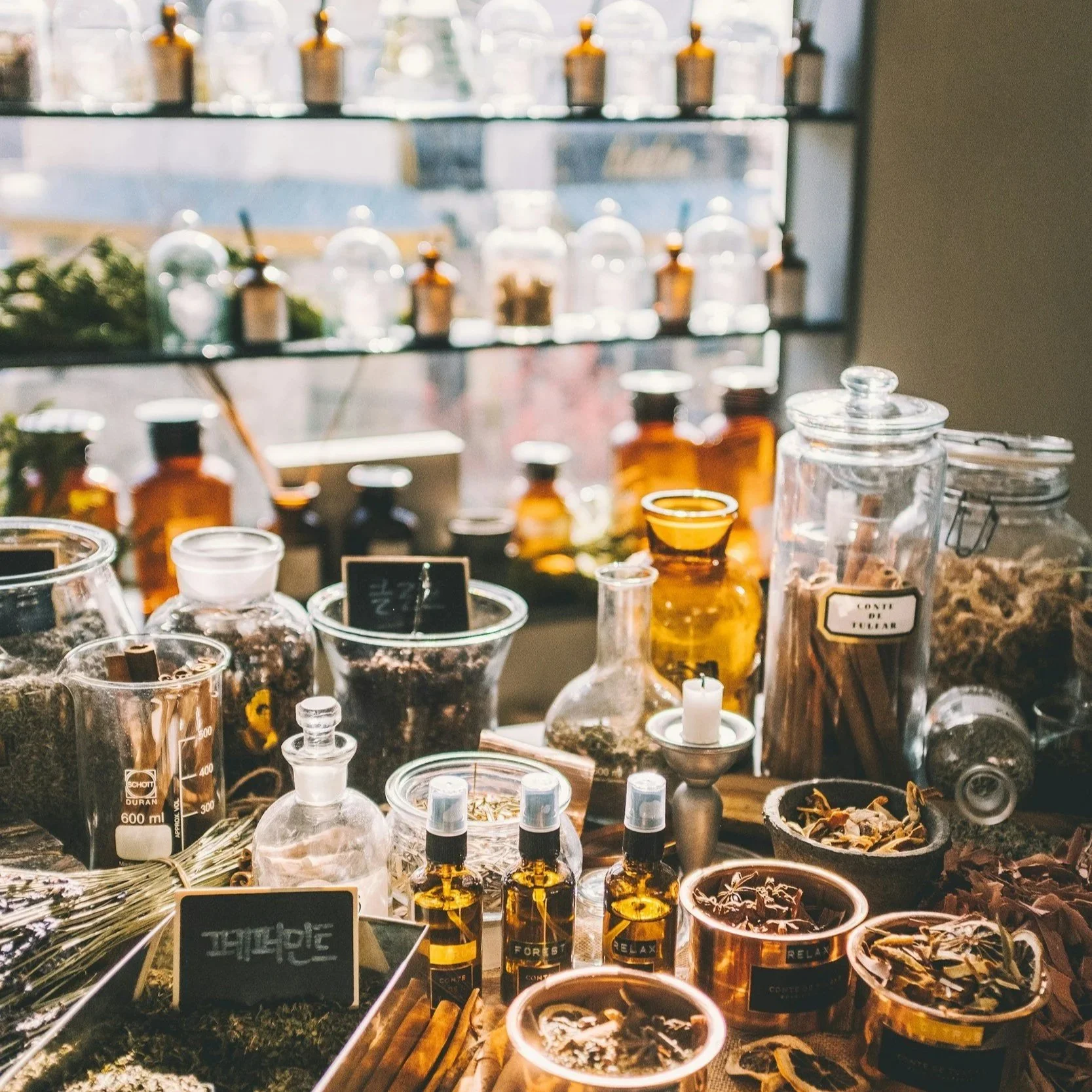 A display of various glass jars and bottles containing herbs, spices, and oils on a wooden table, with shelves of similar jars in the background, illuminated by natural light from a window.