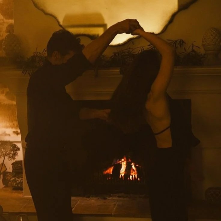 Two people dancing together in front of a fireplace, their silhouettes framed by warm light in an intimate and timeless moment.