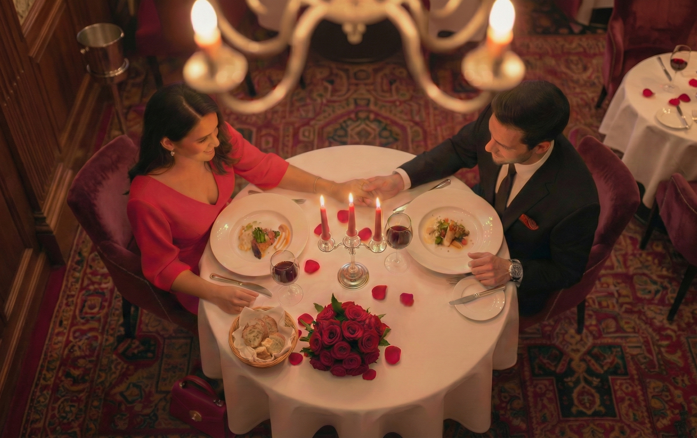 An intimate dinner for two seen from above, with candlelight, roses, and hands gently held across a table set for a romantic evening.