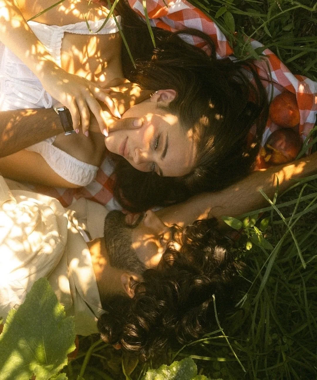 A couple lying together on a checkered blanket in a garden, surrounded by greenery and dappled sunlight, sharing a quiet, intimate moment.