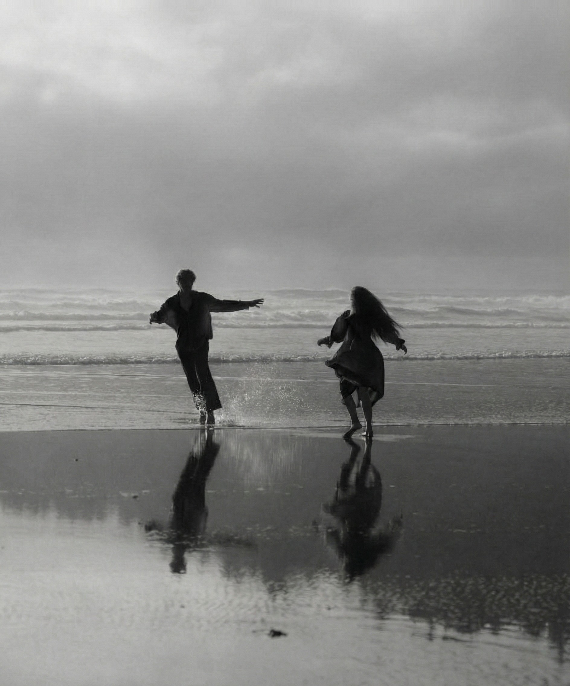 Two people running together along the shoreline, their reflections mirrored on wet sand beneath a soft, overcast sky, capturing a moment of freedom and connection.