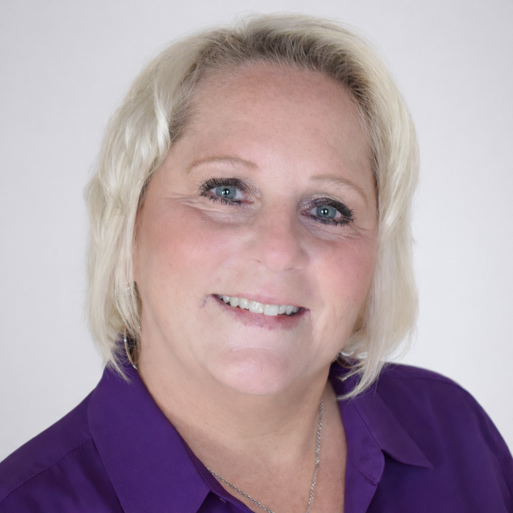 A smiling middle-aged woman with blonde hair and blue eyes, wearing a purple shirt, against a plain white background.