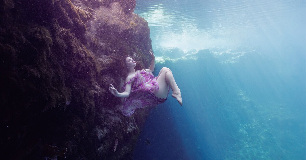 A woman swimming underwater near rocks, wearing a purple dress.